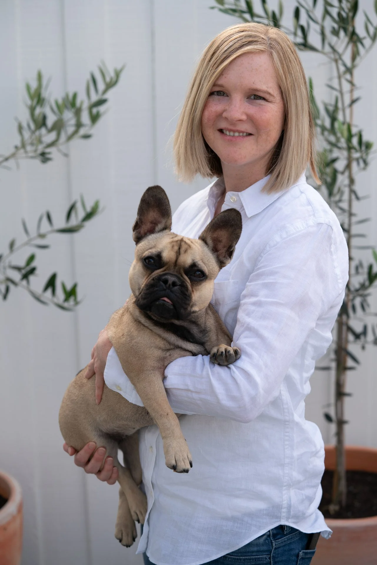 A woman with blonde hair dressed in a white shirt holding a French Bulldog puppy outdoors.