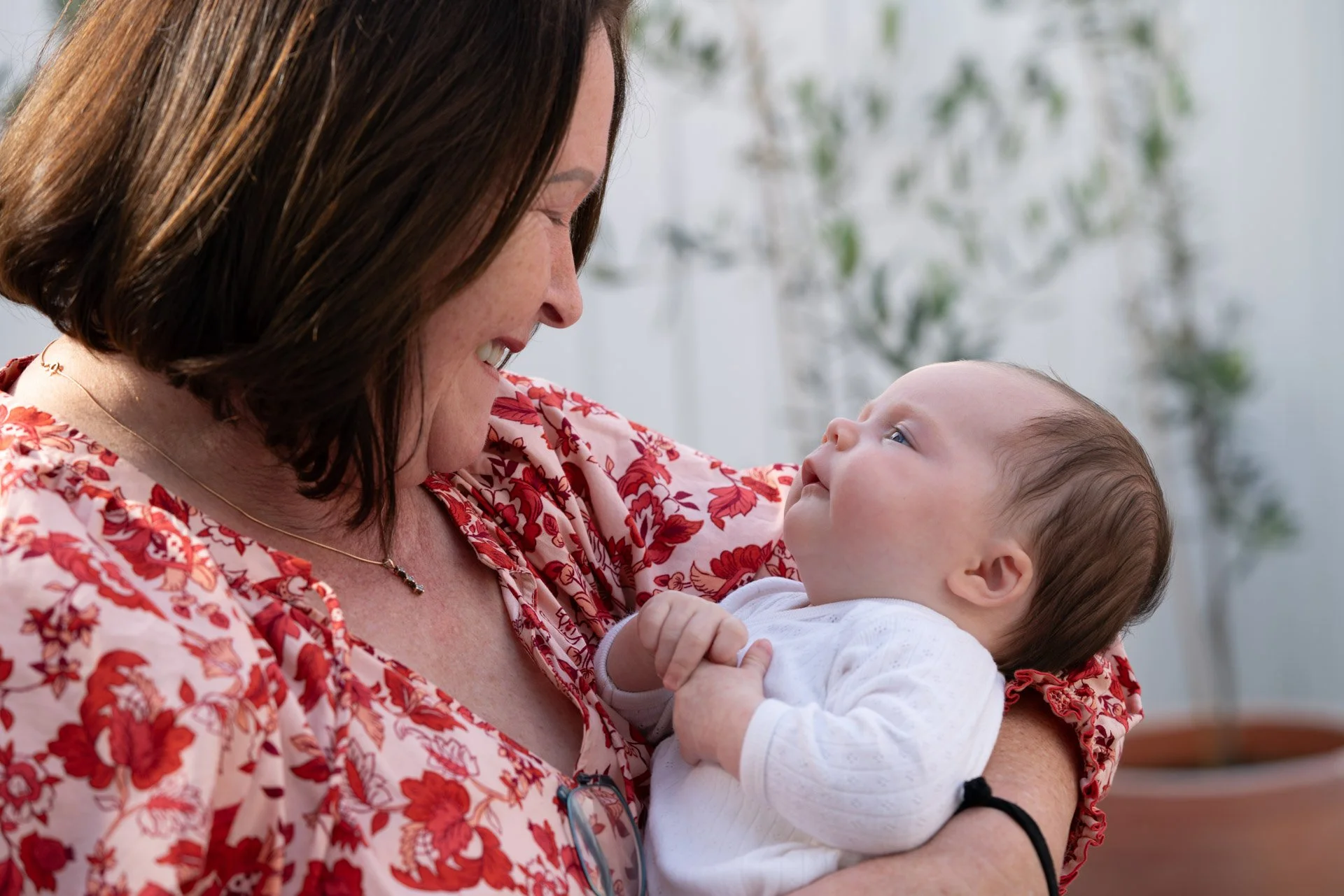 A woman with brown hair, wearing a floral blouse, holding a baby and smiling at the baby, who is looking at her.