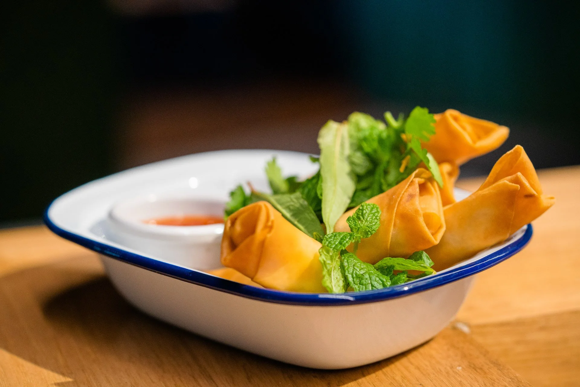 A white bowl with blue rim containing four spring rolls, a side of green lettuce and herbs, and a small dish of dipping sauce, placed on a wooden surface.
