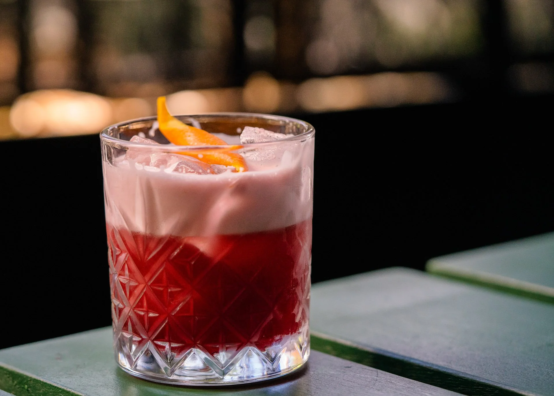 A glass of red cocktail topped with pink foam and garnished with a twist of orange peel, on a green and blue table, with a blurred outdoor background.