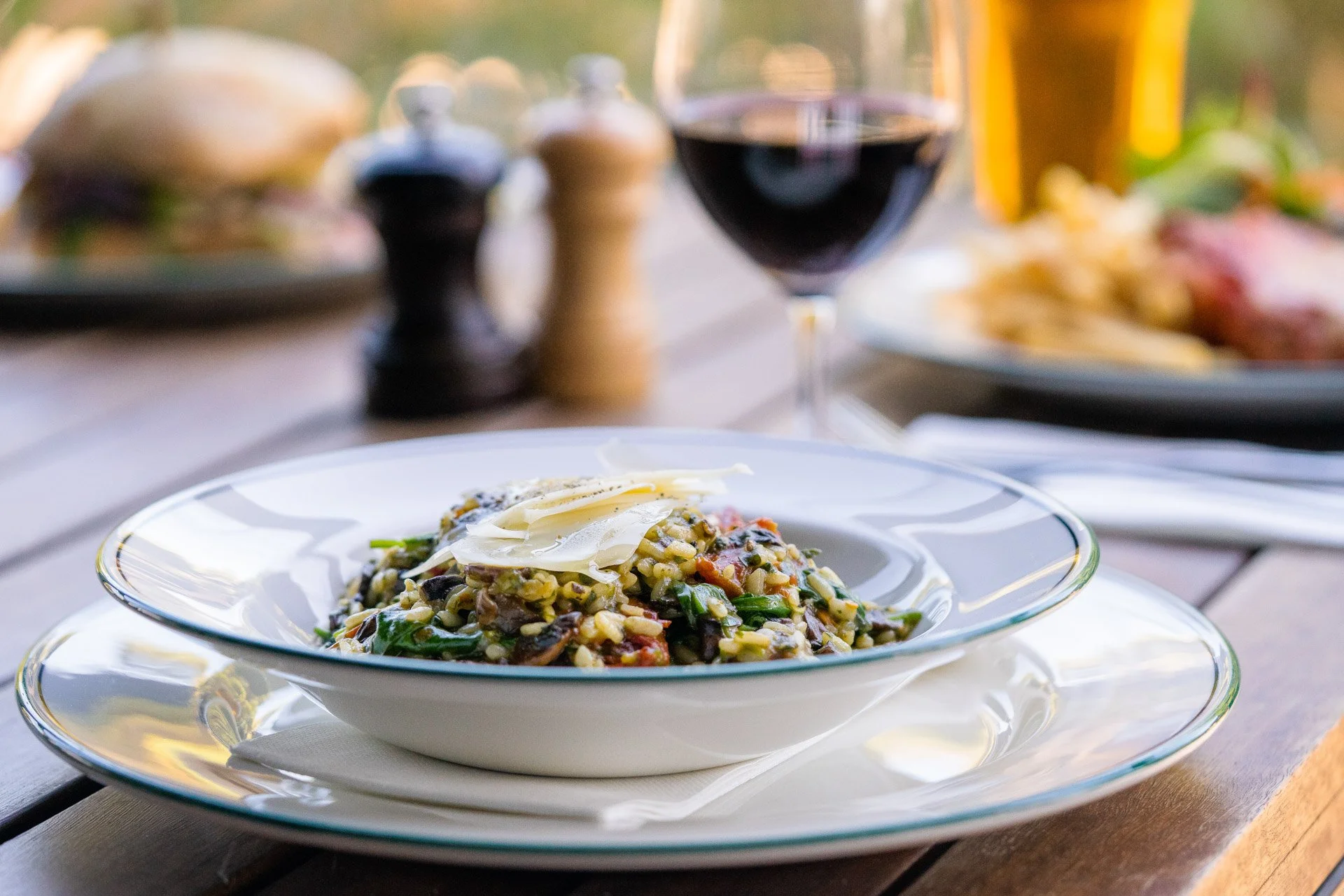 A plate of risotto topped with cheese on a white dish, with a glass of red wine and a beer in the background, set on a wooden table outdoors.