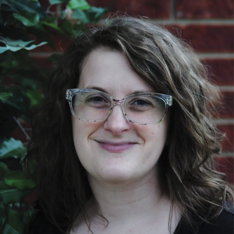 A woman with curly brown hair wearing glasses, smiling outdoors with green foliage and a brick wall in the background.