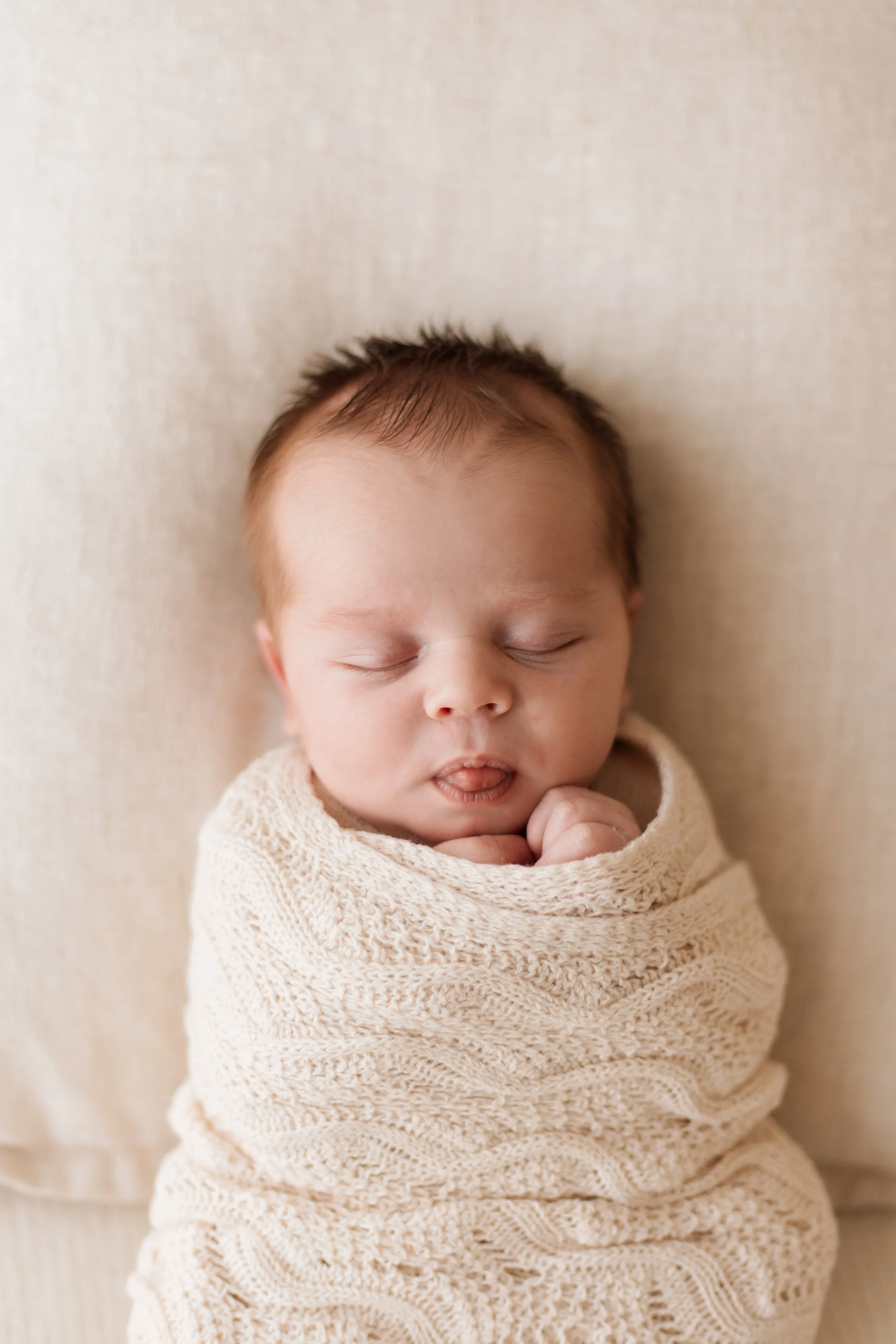 A sleeping baby with closed eyes, wrapped in a cozy beige knit blanket, and lying against a matching background.
