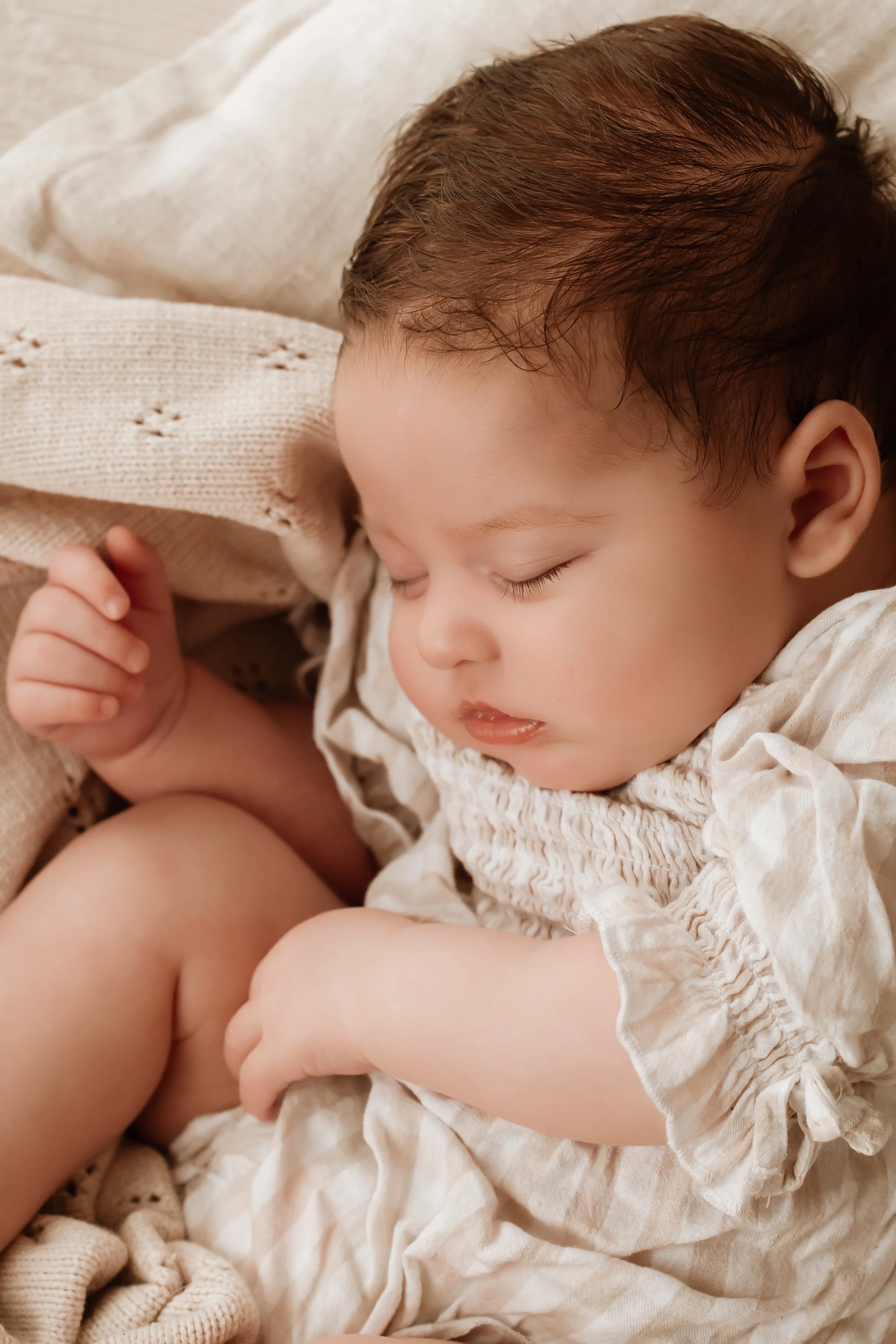 A young child sleeping peacefully with eyes closed, wearing a light-colored outfit, surrounded by soft blankets and knitted items.