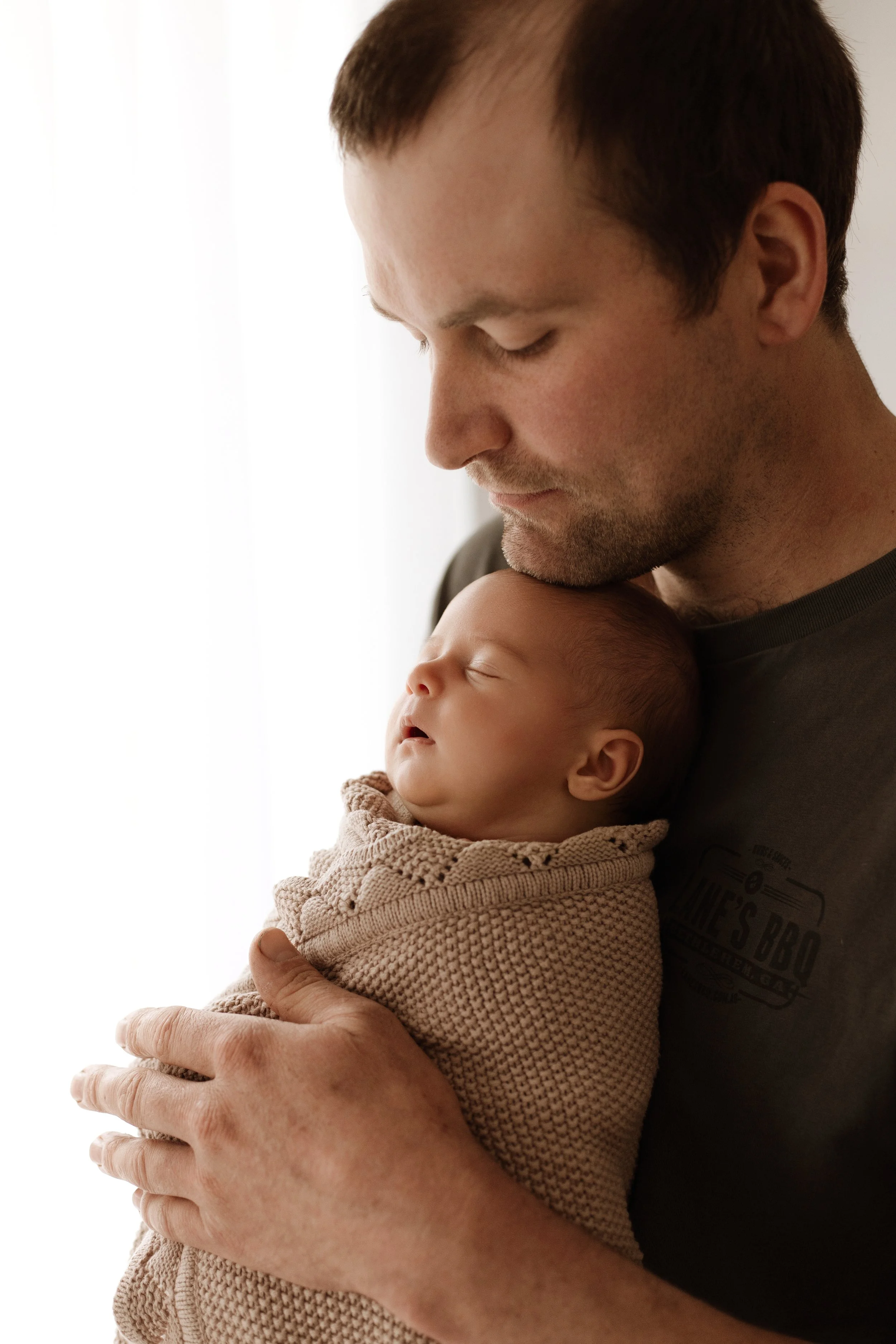 A man holding a sleeping baby wrapped in a beige knitted blanket, close to his chest, with a calm, tender expression.