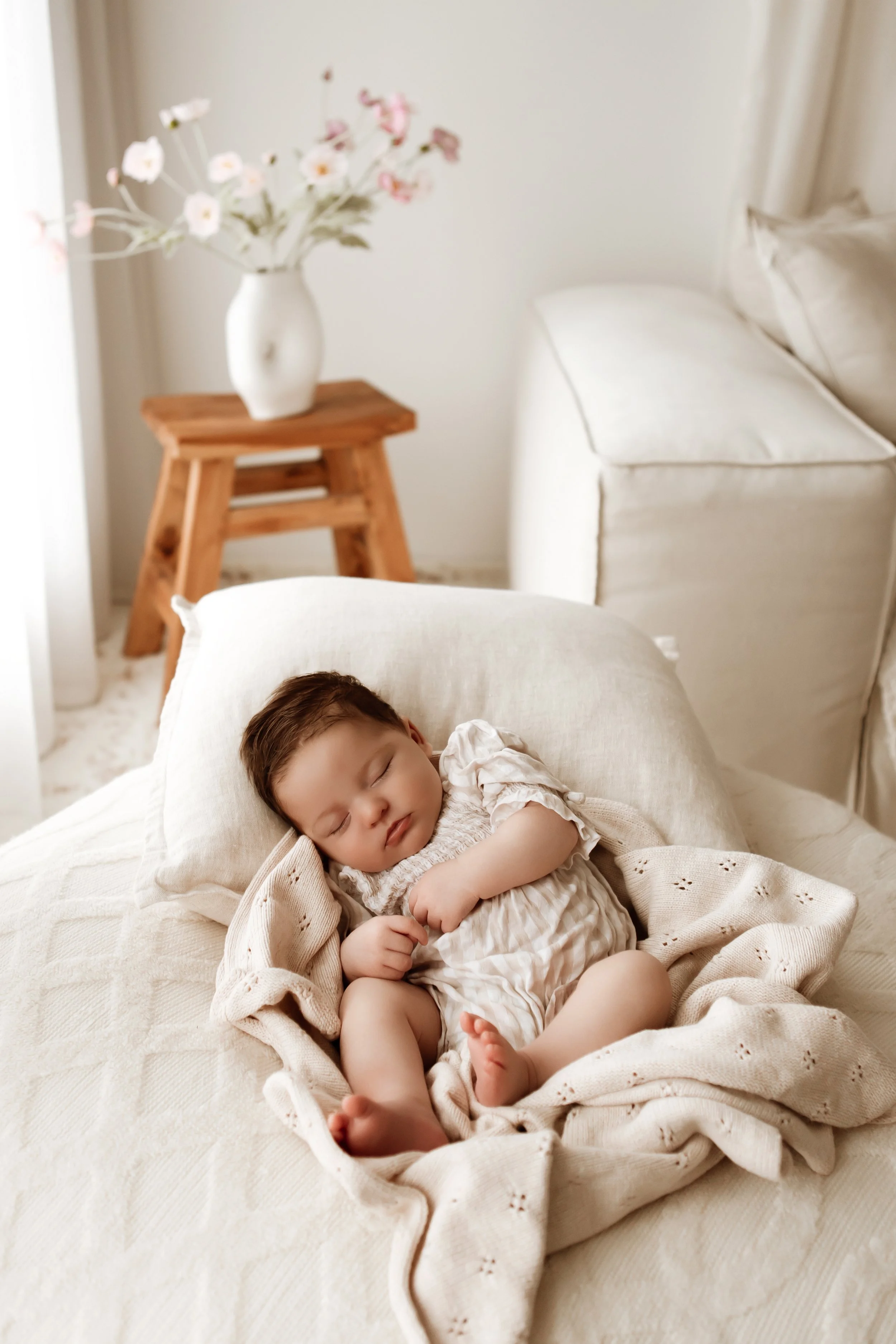 A young child with brown hair sleeping peacefully on a cream-colored bed, covered with a matching blanket, in a bedroom with white walls, a wooden side table with a white vase of pink and white flowers, and a white armchair.