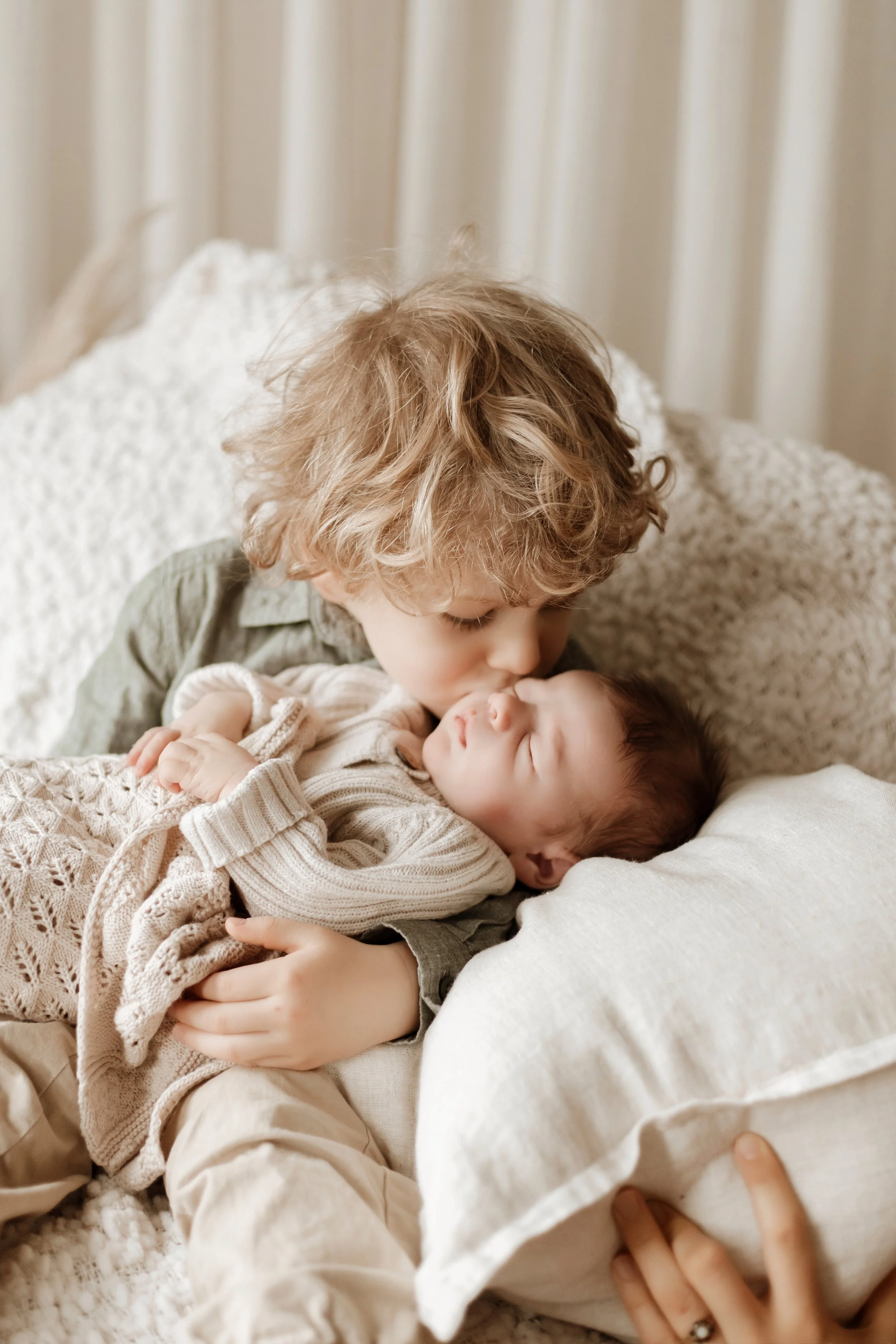A young boy with curly blond hair and a beige shirt kisses a sleeping baby on the forehead while lying on a bed with beige bedding and white pillows.