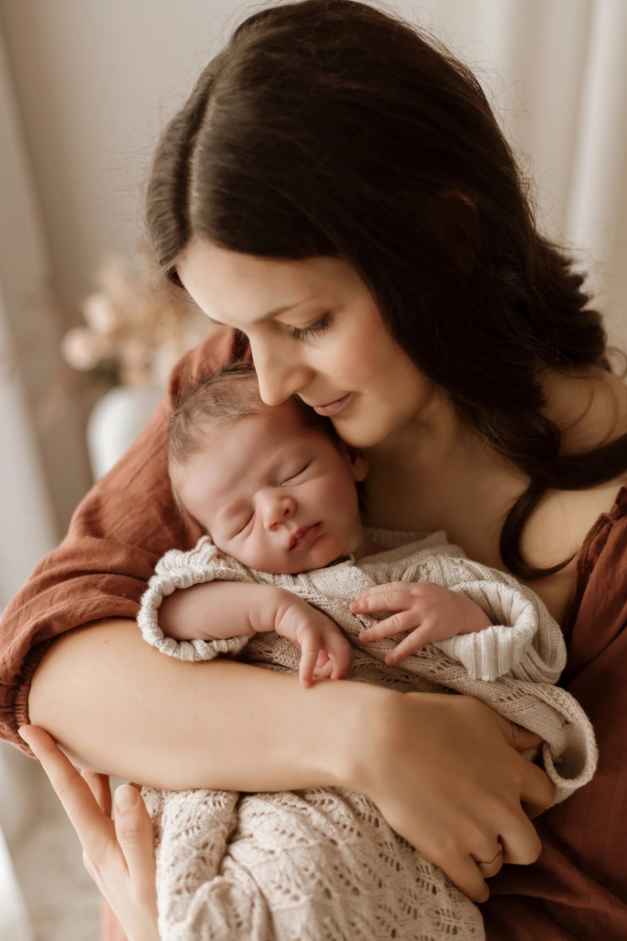 A woman holding a sleeping baby close to her face, with their eyes closed, in a warm indoor setting.