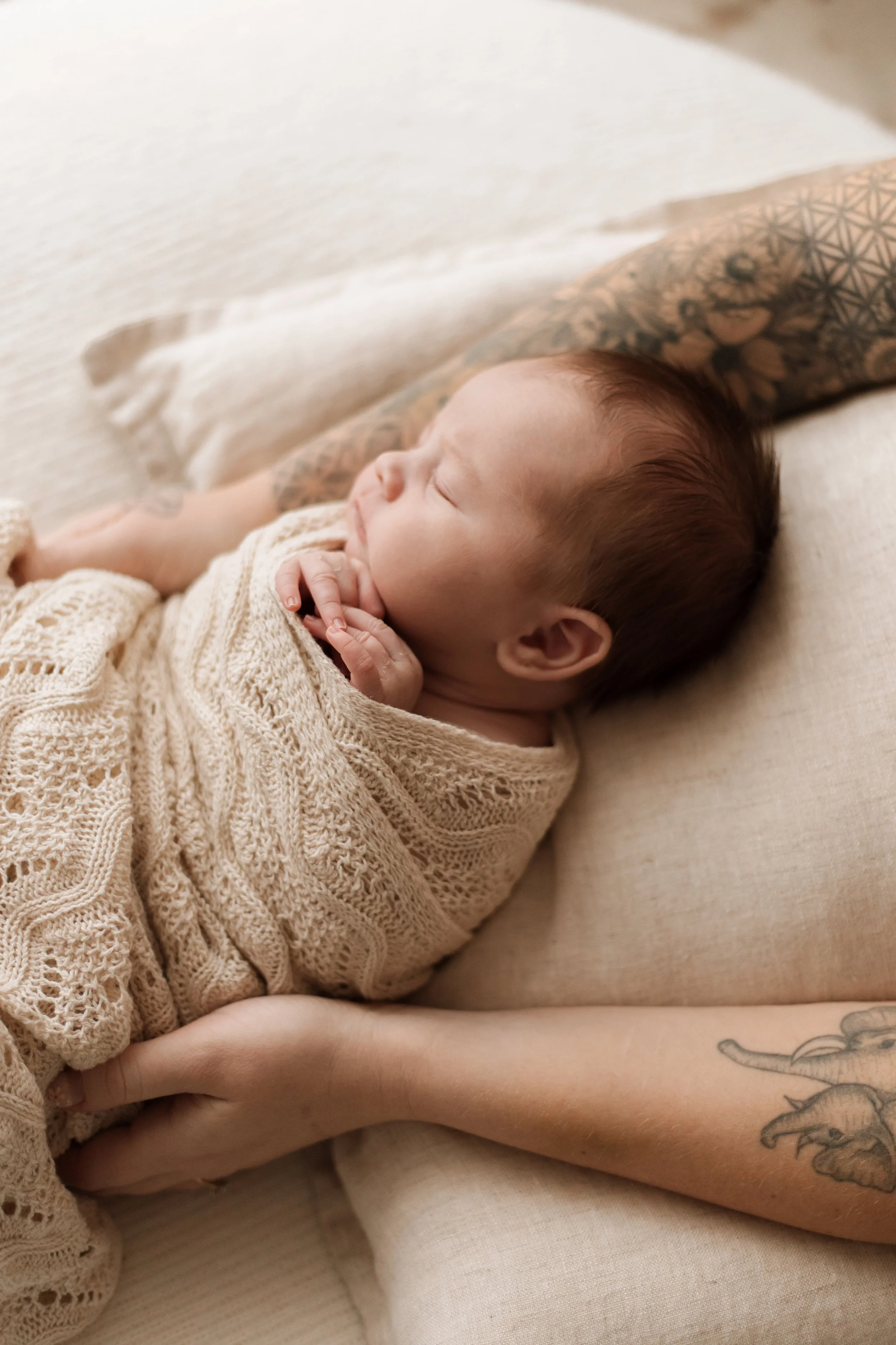 A sleeping baby wrapped in a beige knitted blanket, resting on a cream-colored pillow, with a tattooed arm visible.