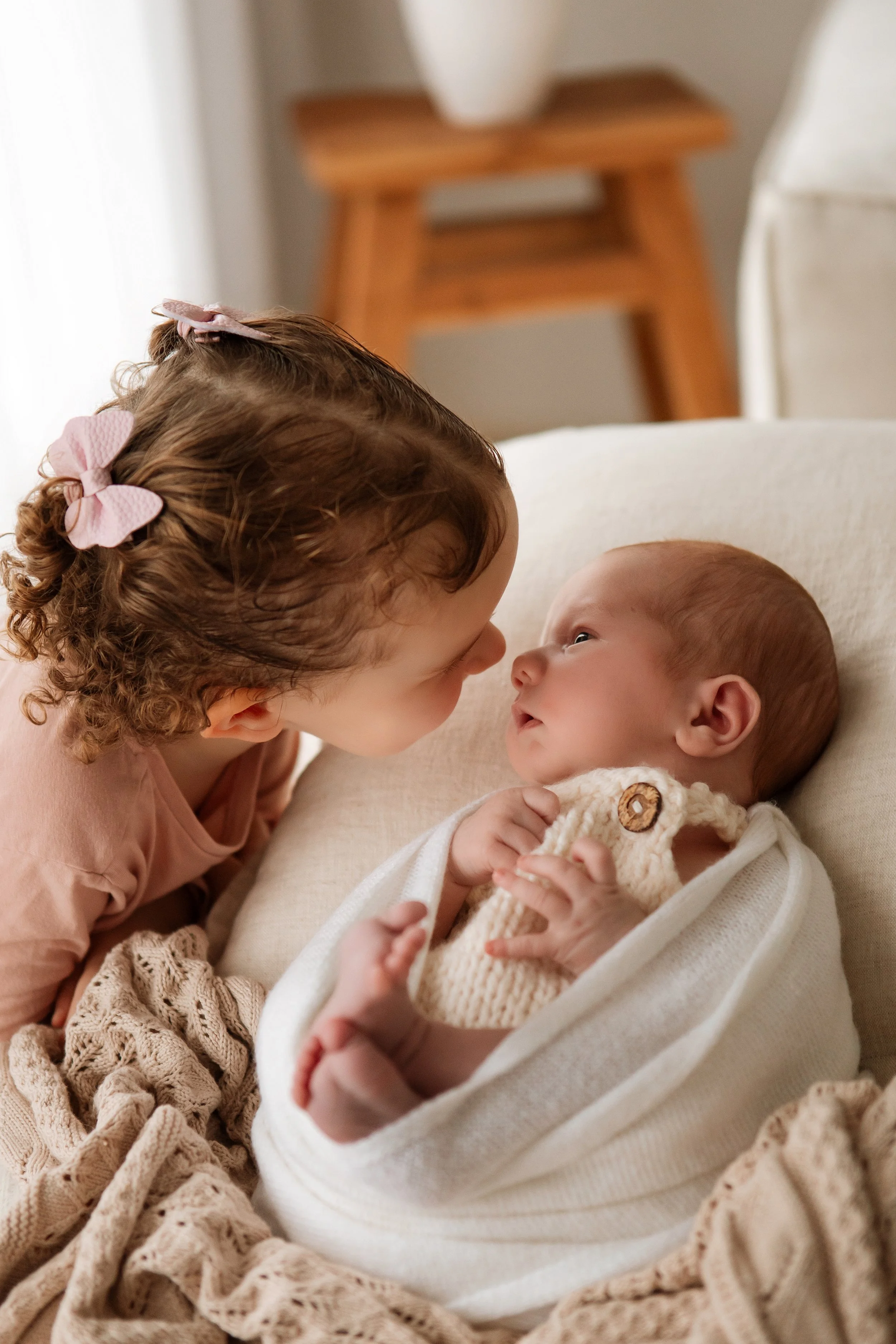 A young girl with curly hair and pink hair bows leans close to an infant lying in a crib, gazing into each other's eyes.