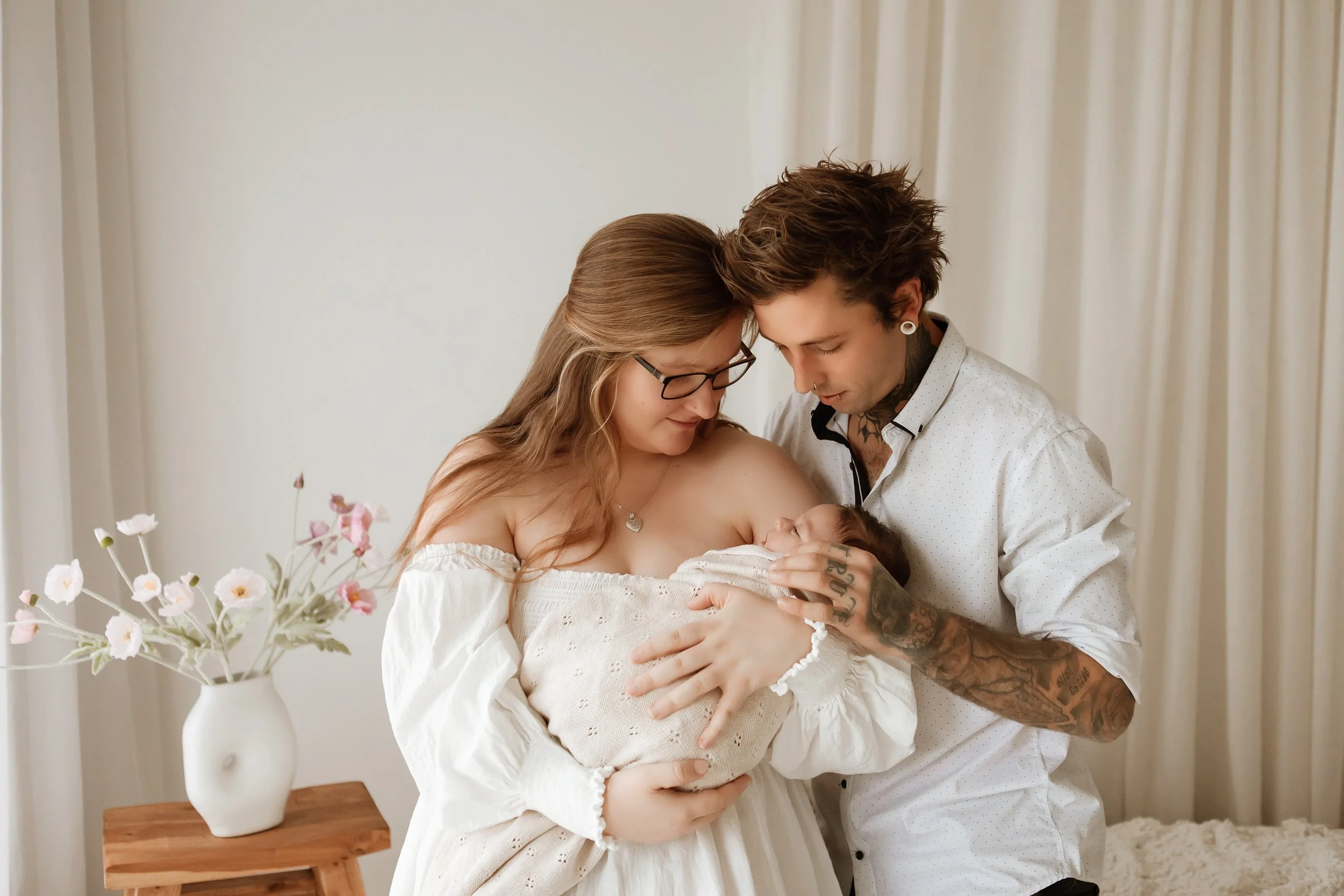 A couple holding their newborn baby in a bright room with cream-colored curtains. The woman has long brown hair, glasses, and is wearing an off-shoulder white dress. The man has short, tousled hair, tattoos, earrings, and is wearing a white shirt. A 