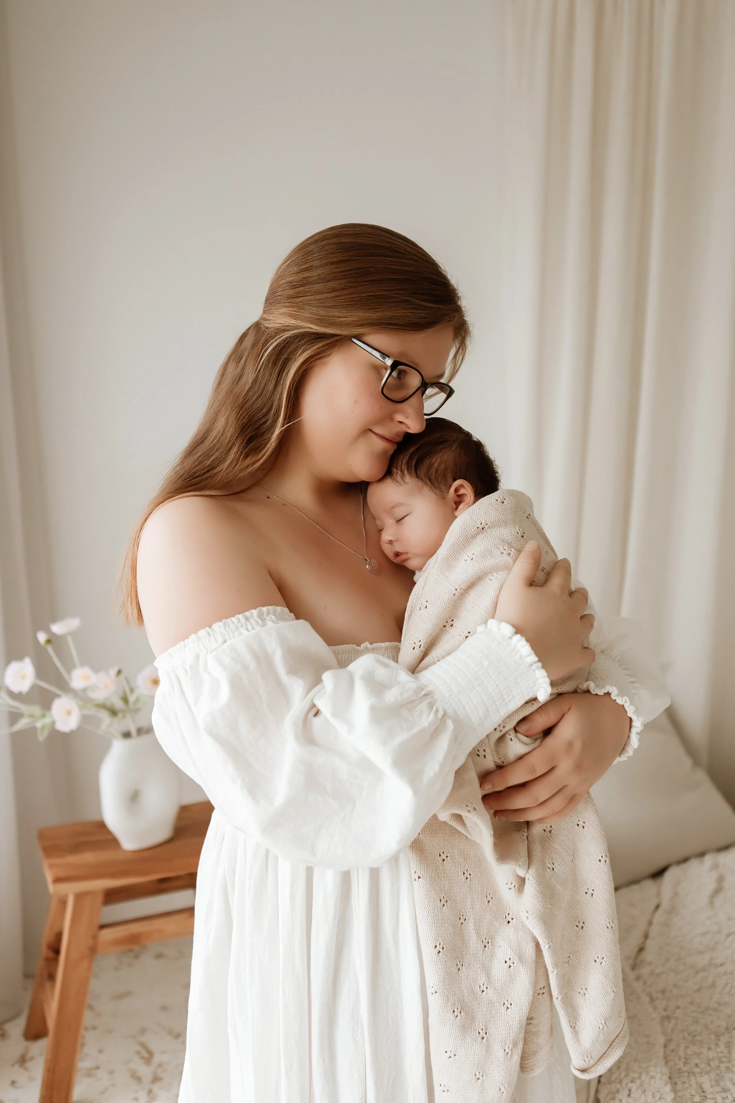 A woman with glasses and long brown hair holding a sleeping baby in a beige outfit. They are indoors, with a vase of white flowers on a wooden table in the background.