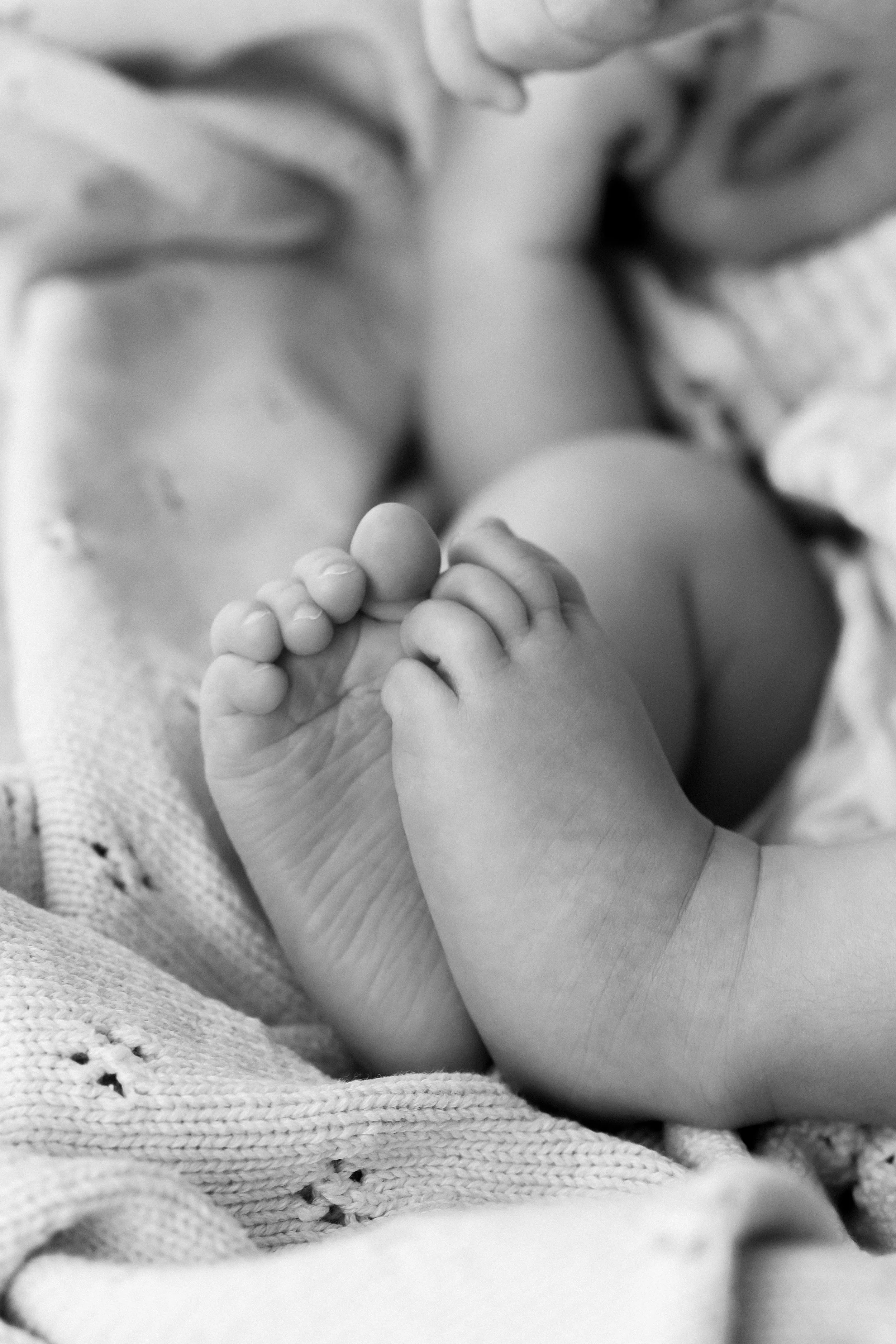Close-up black and white photo of a baby holding an adult's finger, with the baby's face partially visible, lying on a textured blanket.