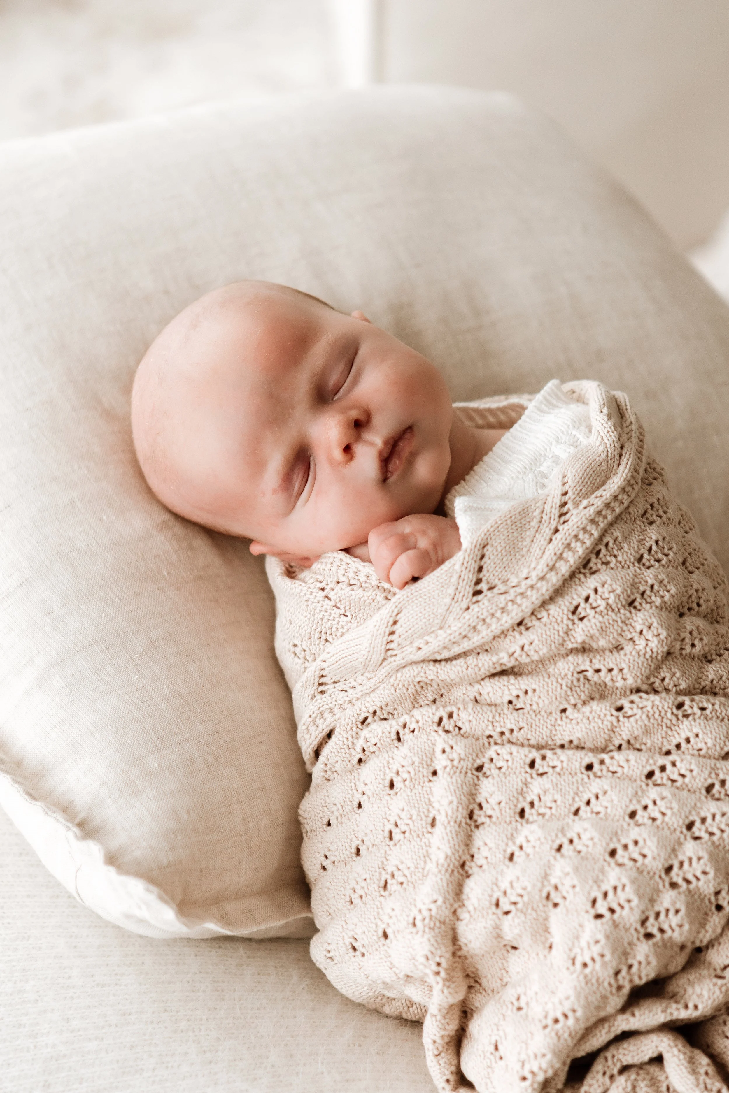 A sleeping baby with a bald head, peacefully resting on a pillow, wrapped in a knitted beige blanket, in a cozy and soft setting.