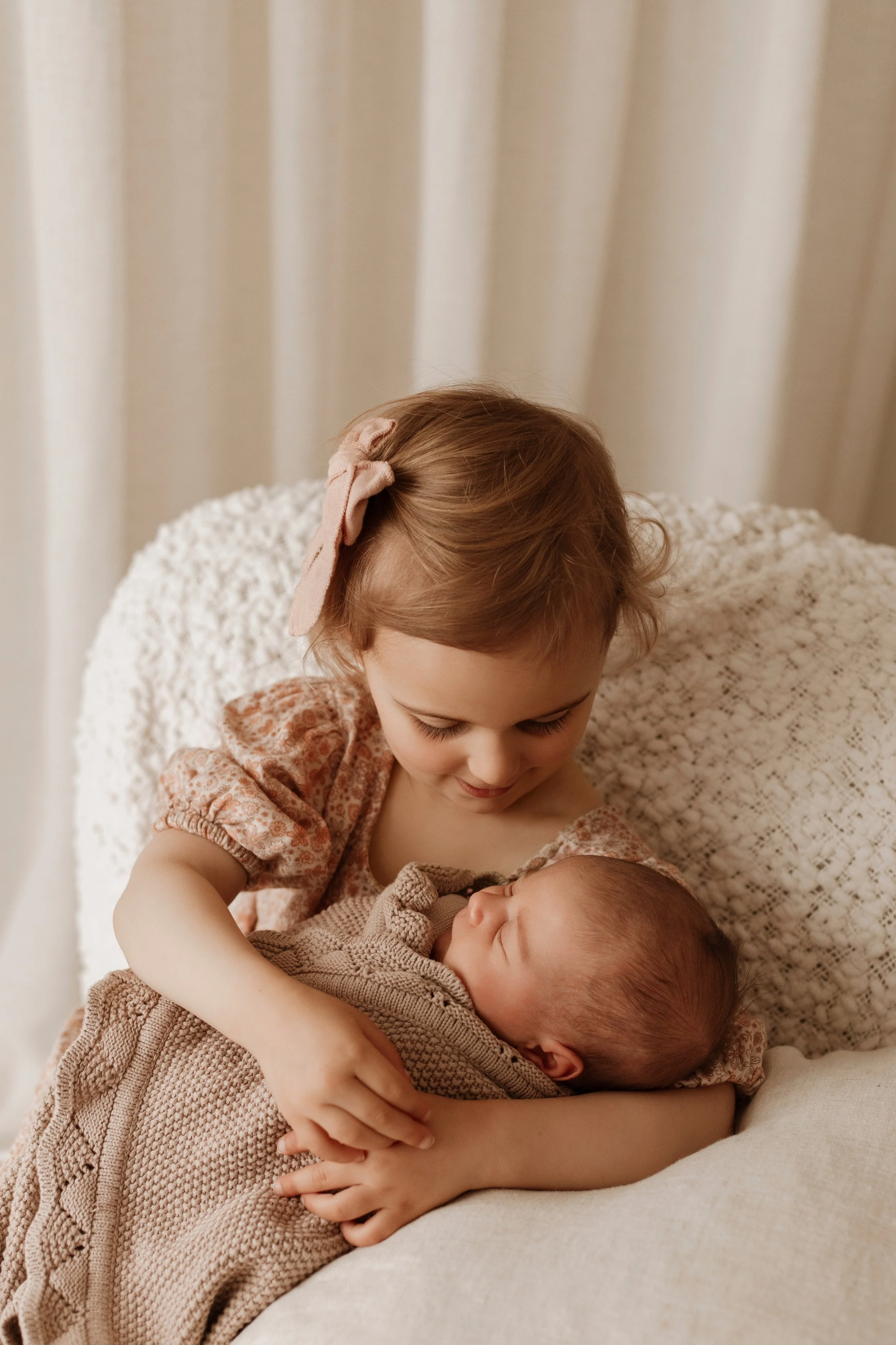 Young girl holding her baby sibling, both smiling softly, in a cozy indoor setting.