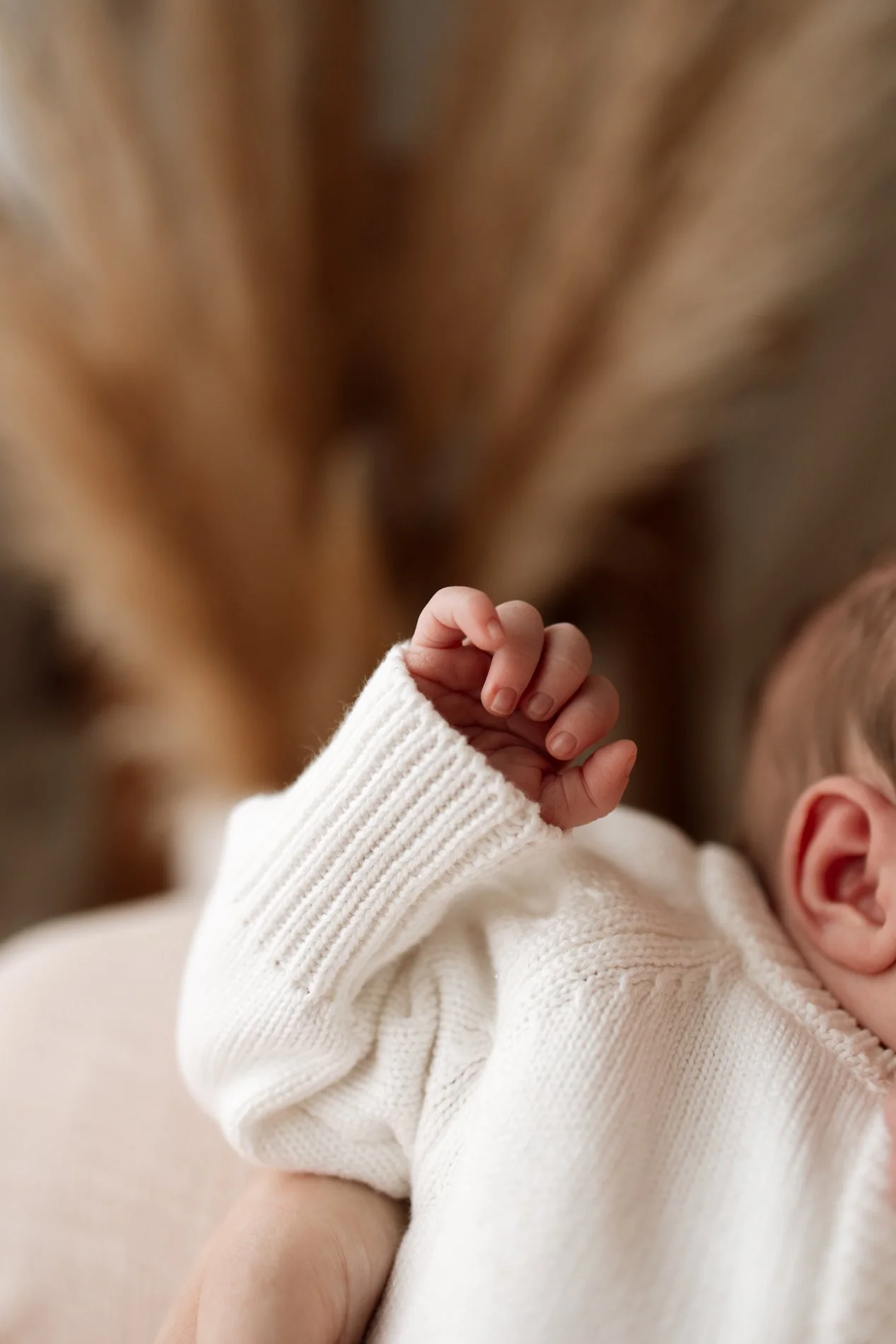 Close-up of a baby's hand in a white knitted sweater resting on an adult's arm, with a blurry background of warm wooden tones.