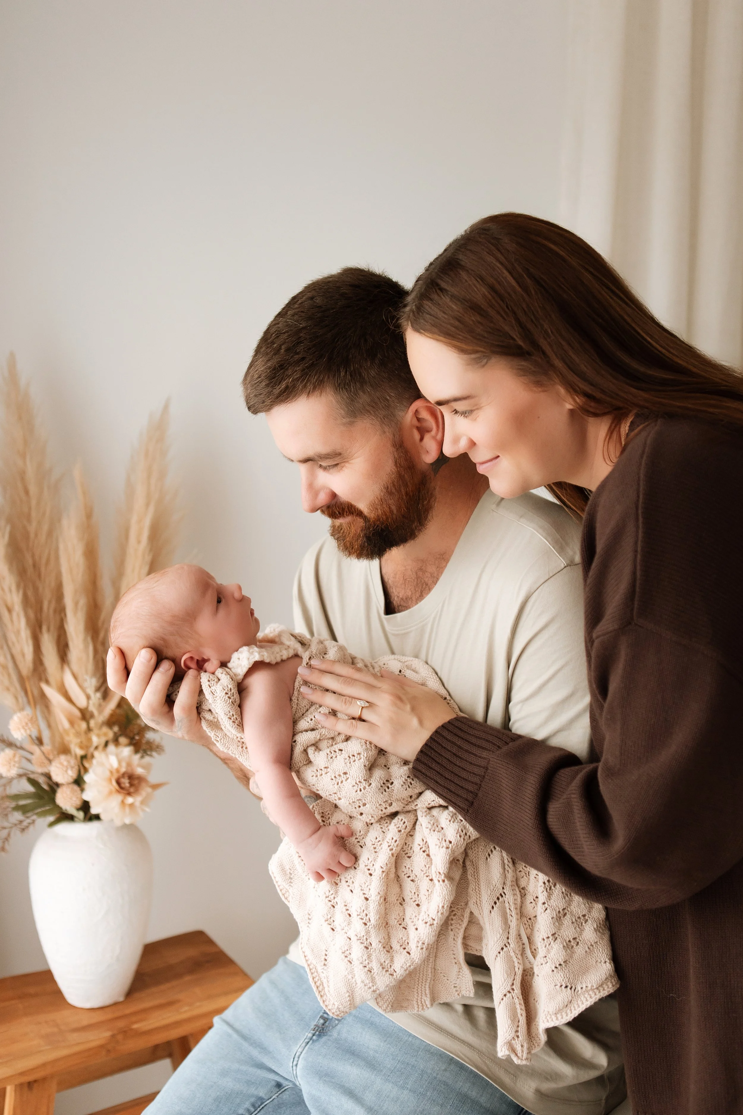 A family with a man, woman, and baby. The man is holding the baby while the woman leans in close to the baby. They are indoors, near a wooden table and a white vase with dried flowers.