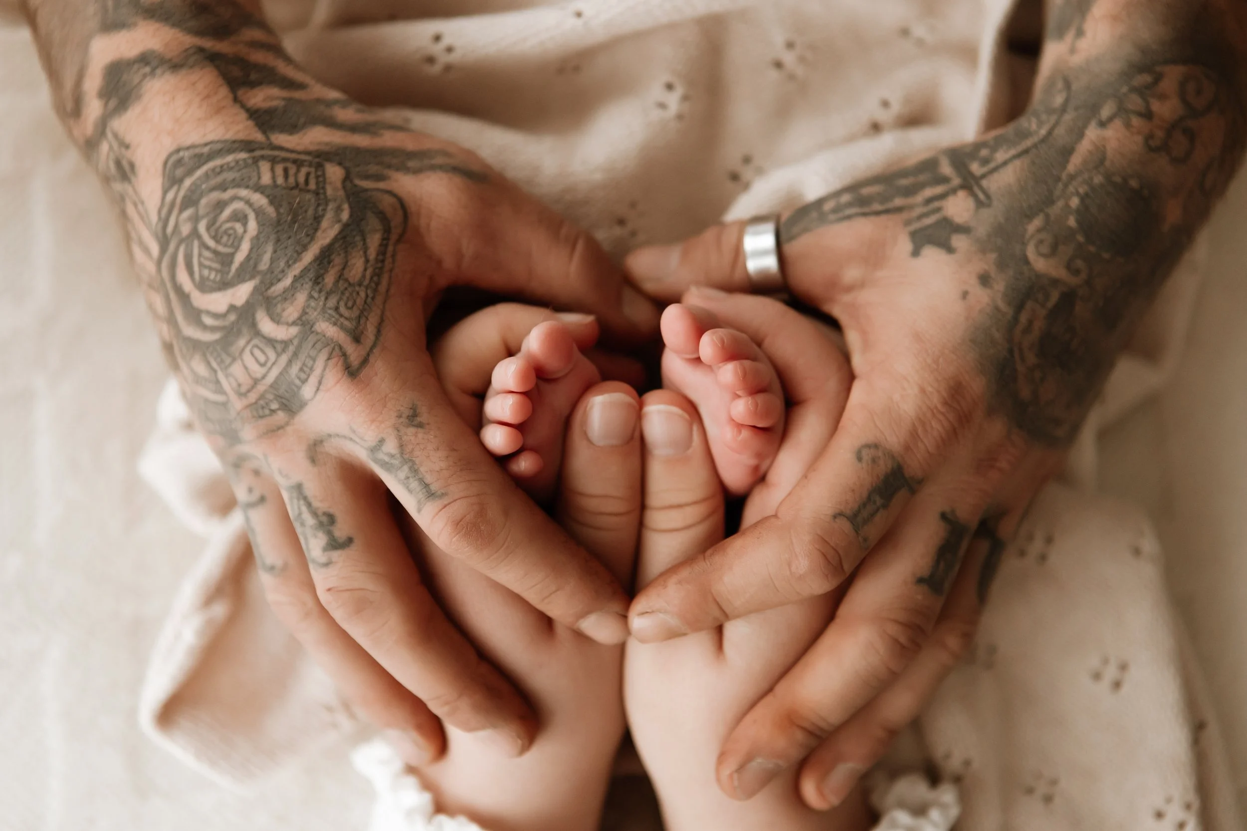 Close-up of a tattooed adult's hands holding a baby's tiny feet, with the adult wearing a silver ring and a cream-colored shirt with small dark dots.