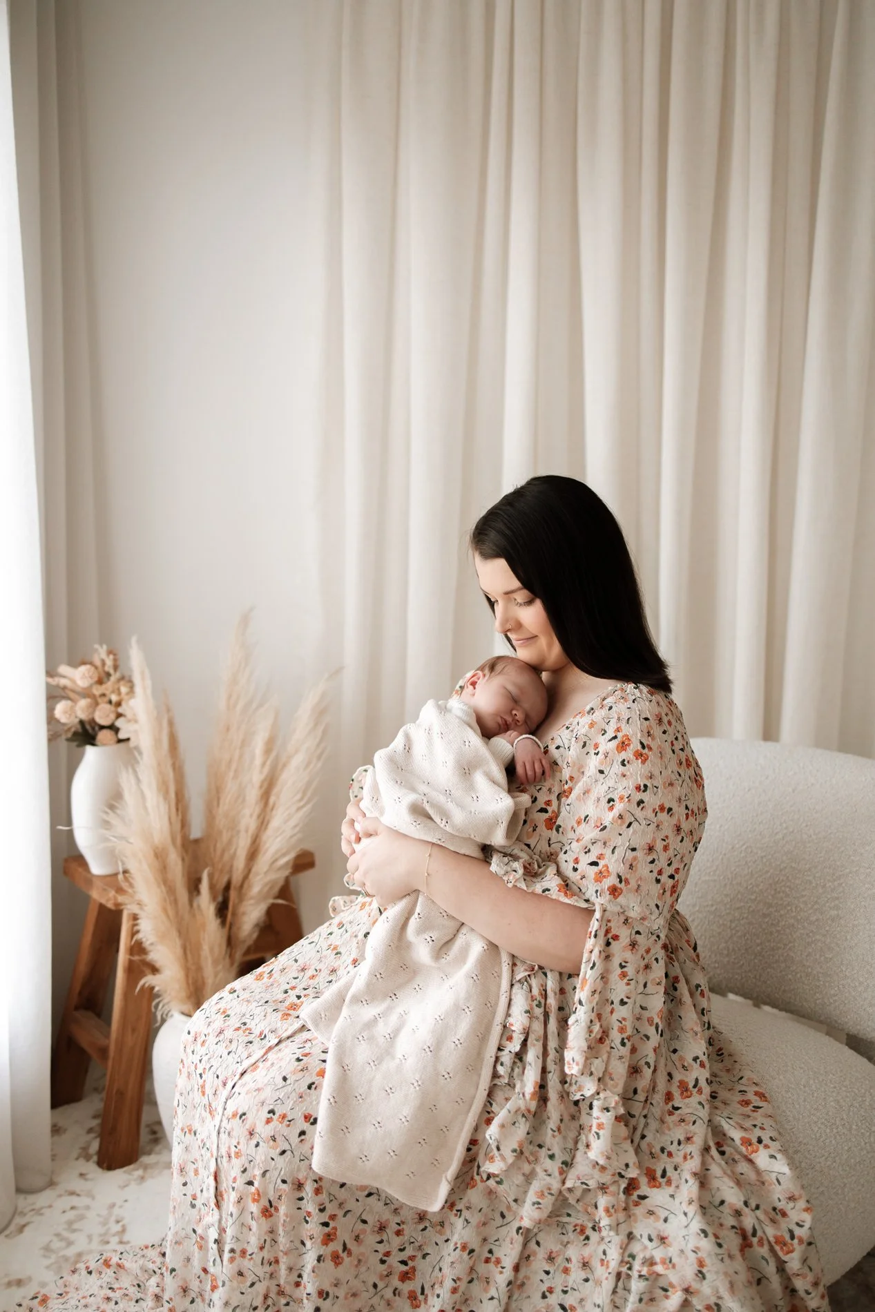 A woman with dark hair holding a sleeping baby on her chest in a cozy, well-lit room with beige curtains and decorative pampas grass.
