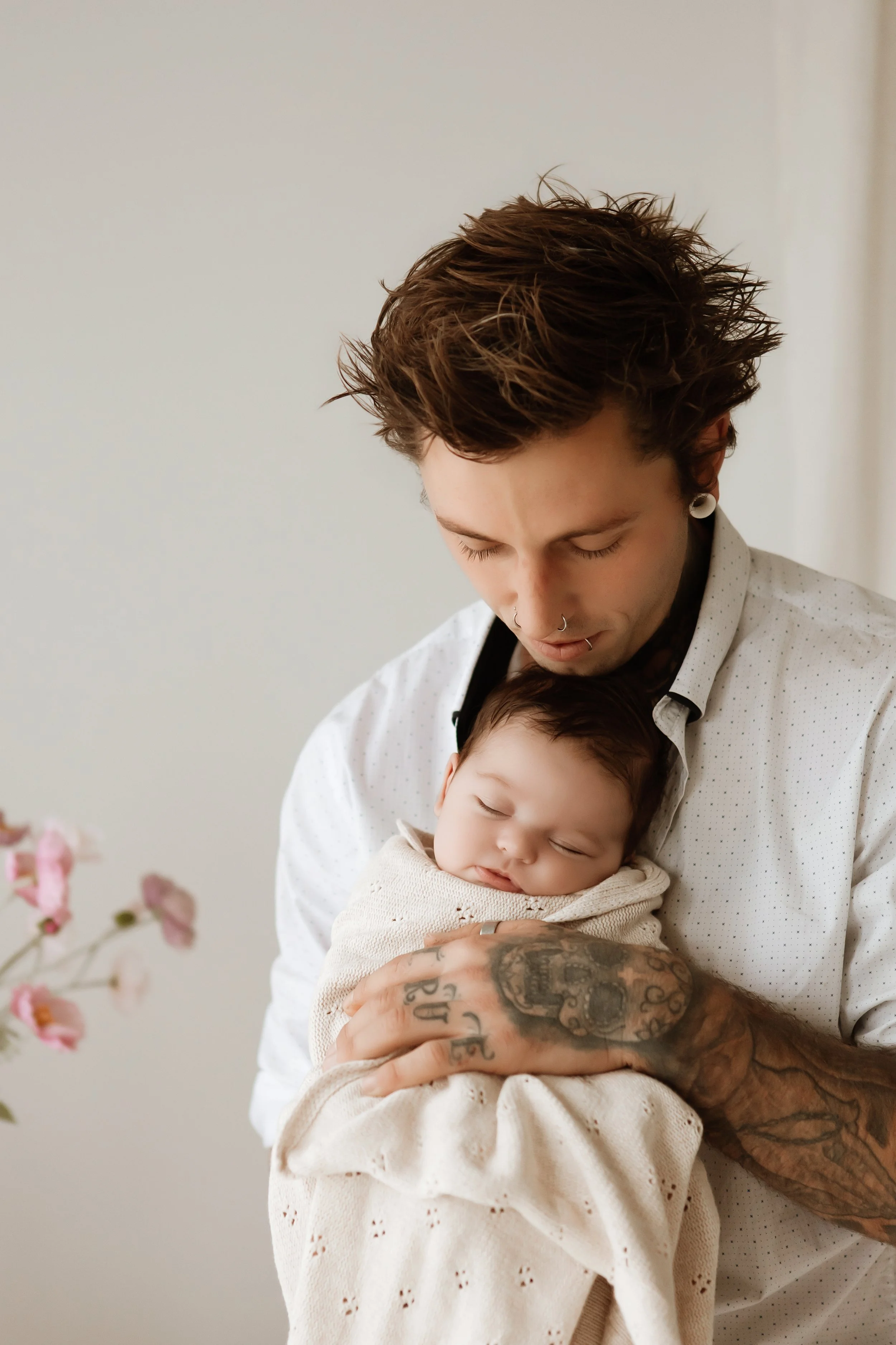 A young man with short, tousled brown hair and tattoos on his arms is holding a sleeping baby wrapped in a cream-colored blanket. The man is wearing a white shirt with small black dots and has gauges in his ears. They are in a room with light-colored