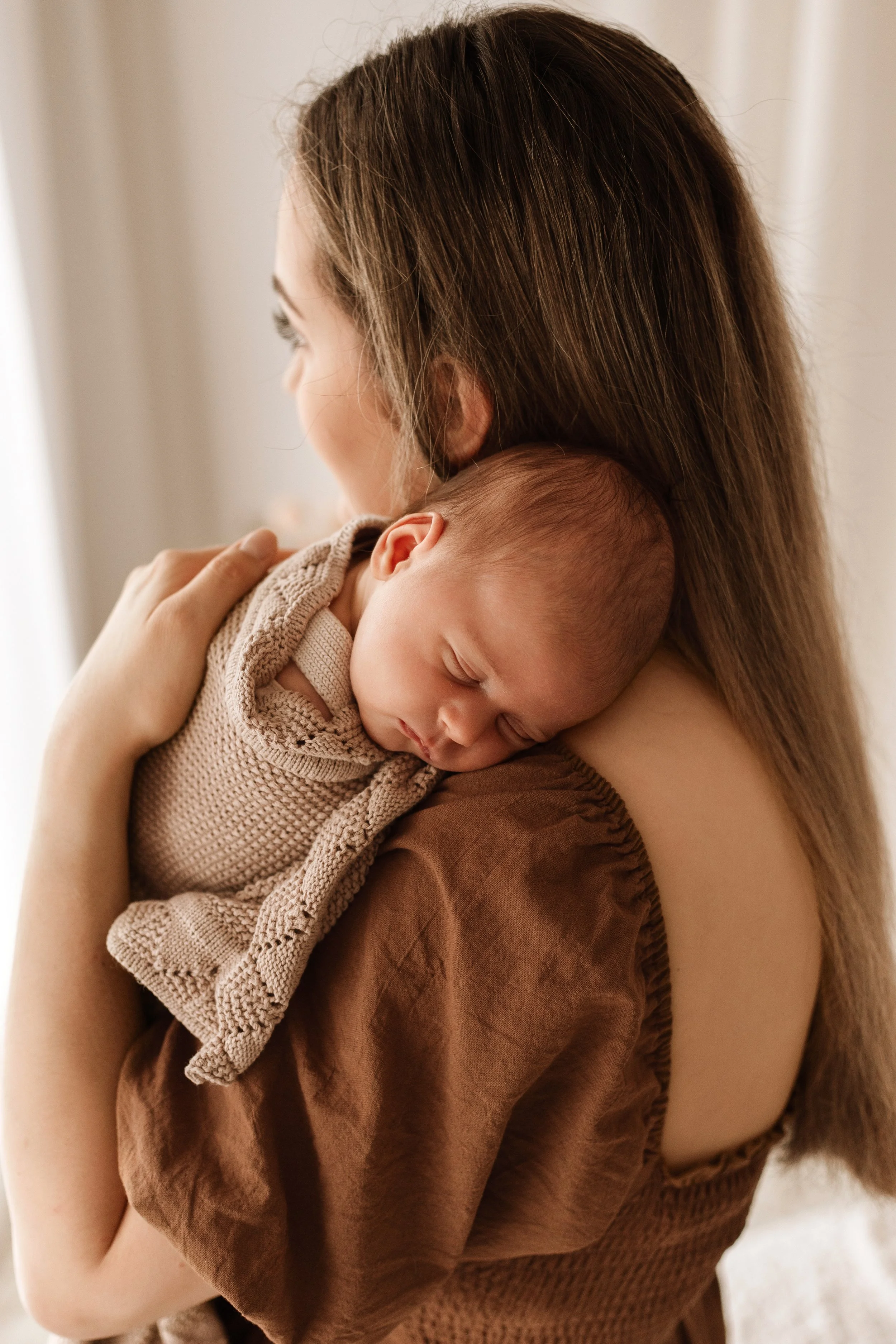 A woman with long brown hair holding a sleeping baby close to her shoulder, seen from the side, in a softly lit room.
