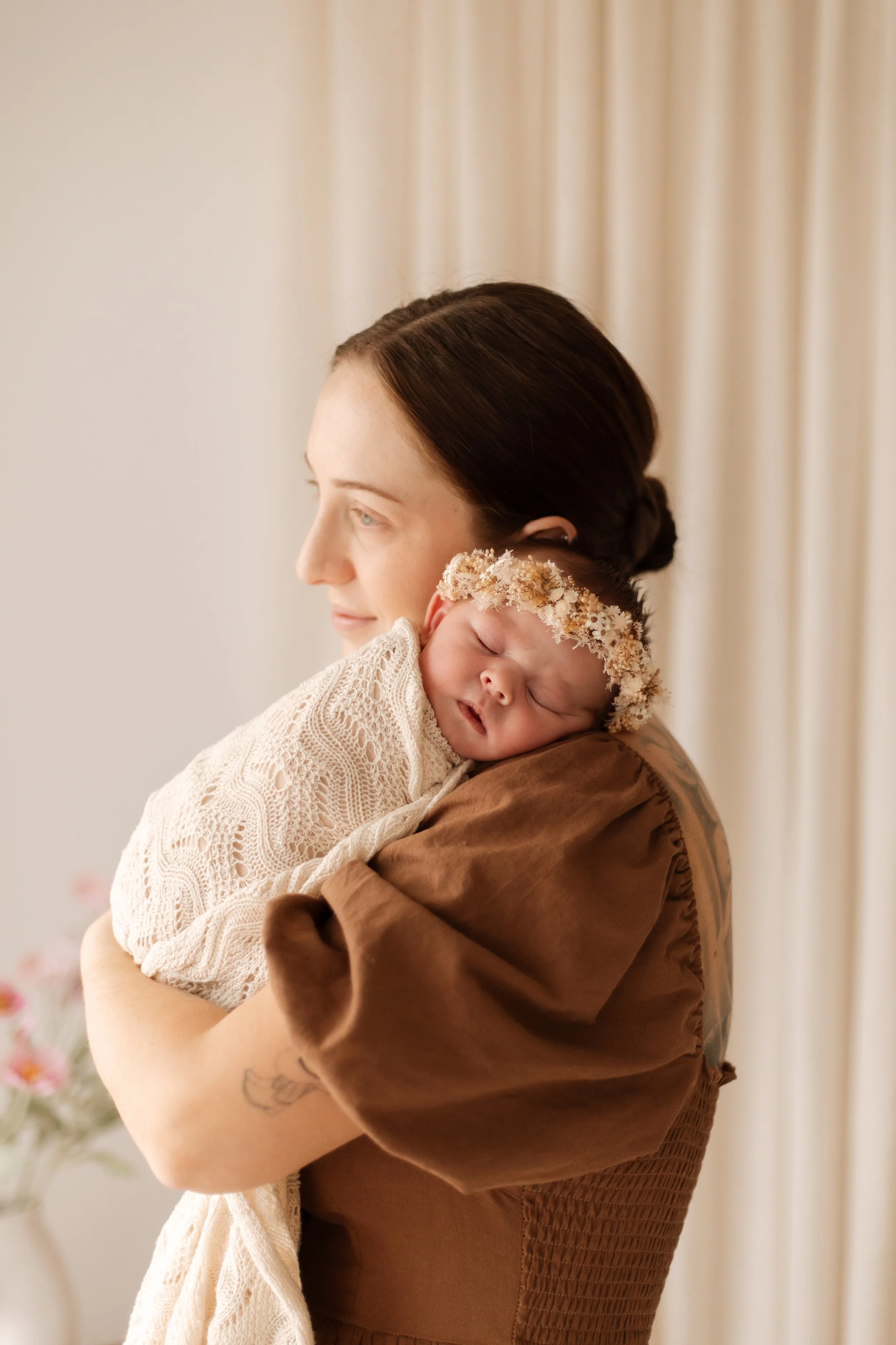 A woman holding a sleeping baby wrapped in a blanket, wearing a flower crown, in a softly lit room with cream-colored curtains and a vase of pink flowers in the background.