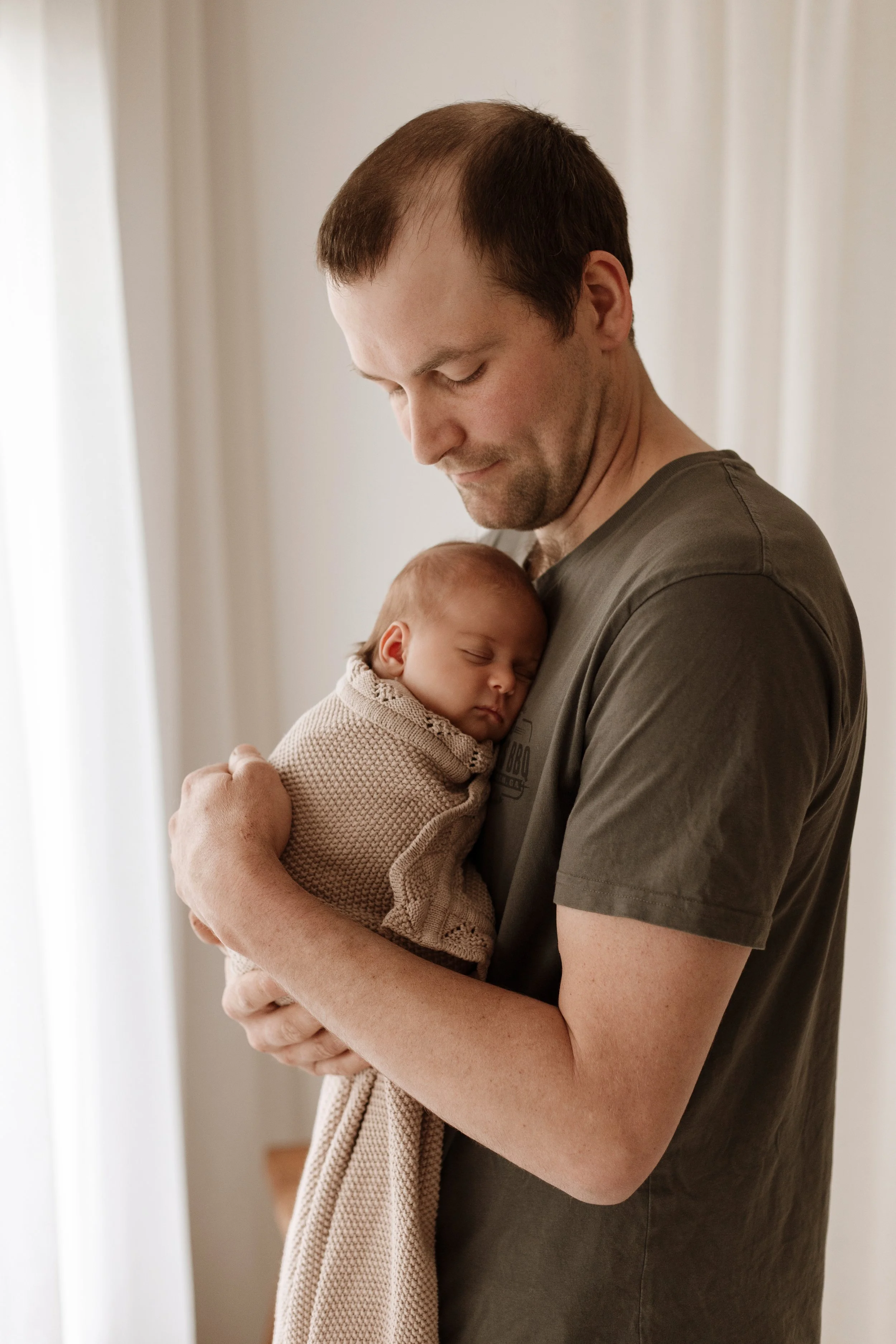 A man holding a sleeping baby wrapped in a blanket, standing near a window with curtains.