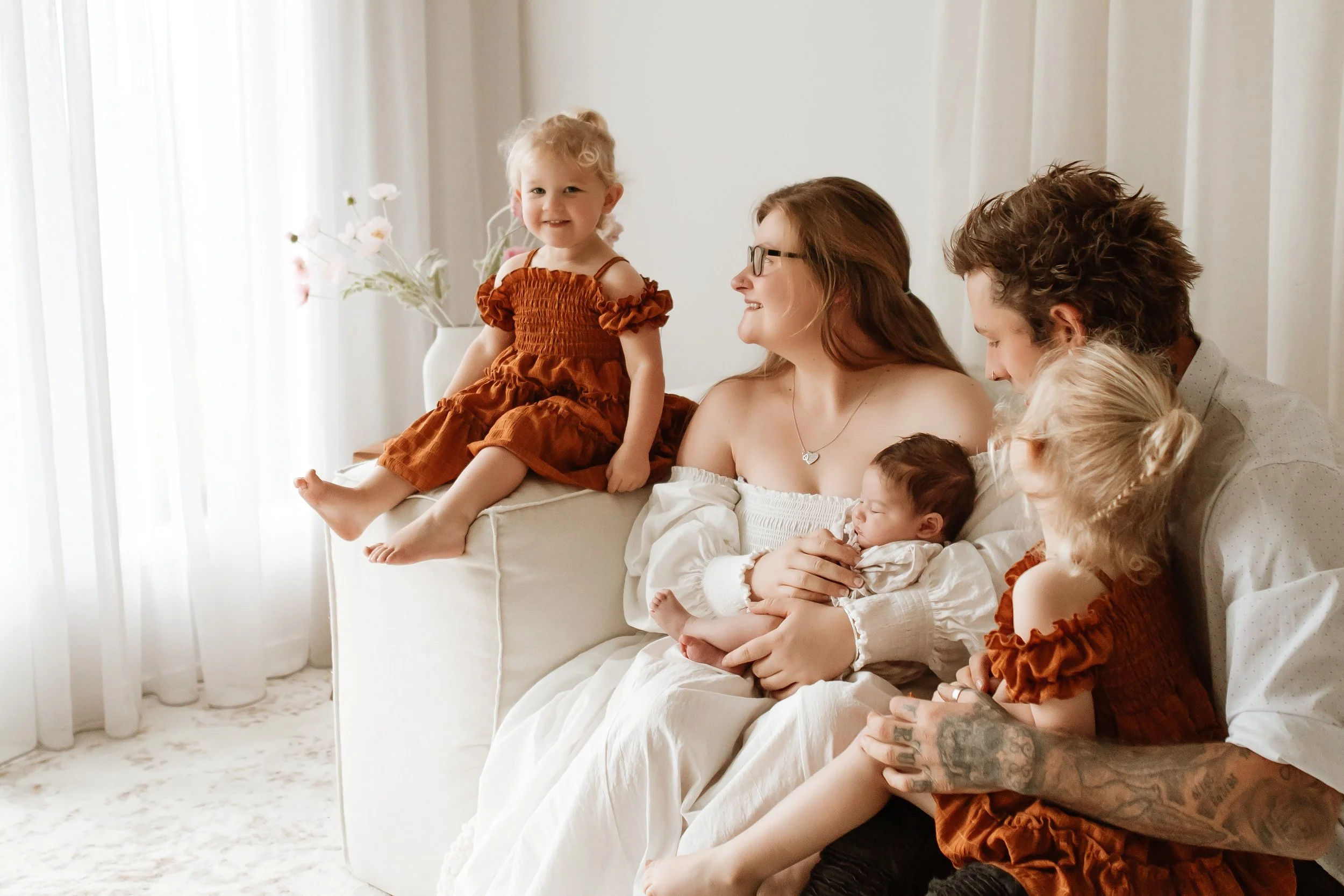 A family sitting on a white couch in a bright room, with a young girl wearing an orange dress sitting on the mother's lap, and other children and a tattooed man nearby.