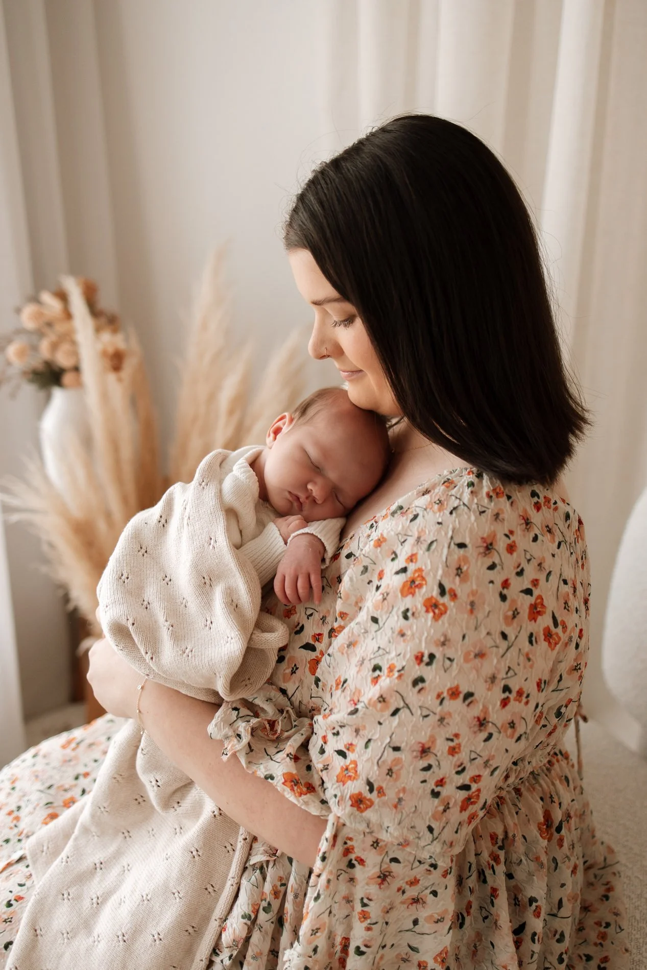 A woman with dark hair holding a sleeping baby in beige clothing in a cozy room with dried flowers and neutral decor.