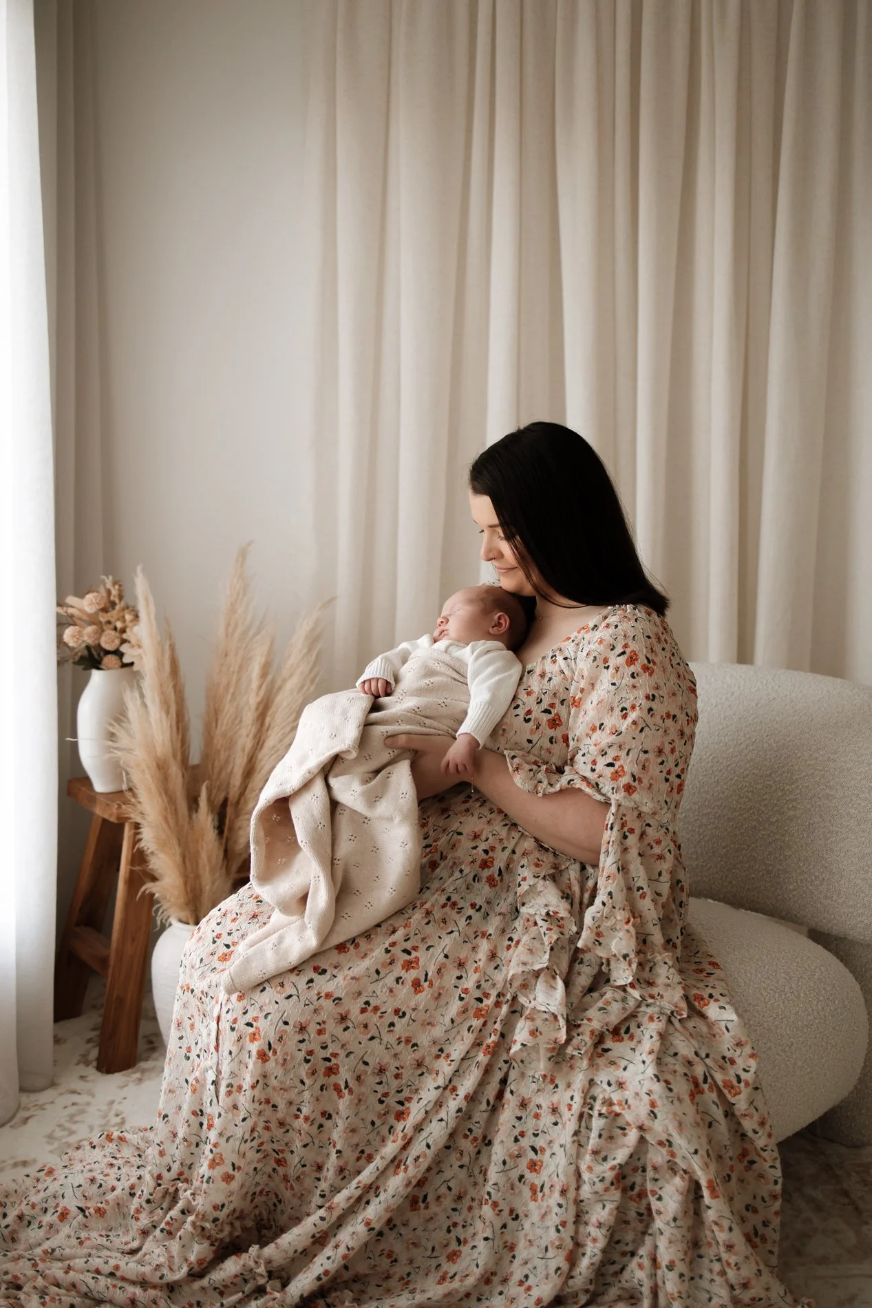 A woman sitting on a beige sofa holding a sleeping baby wrapped in a blanket.
