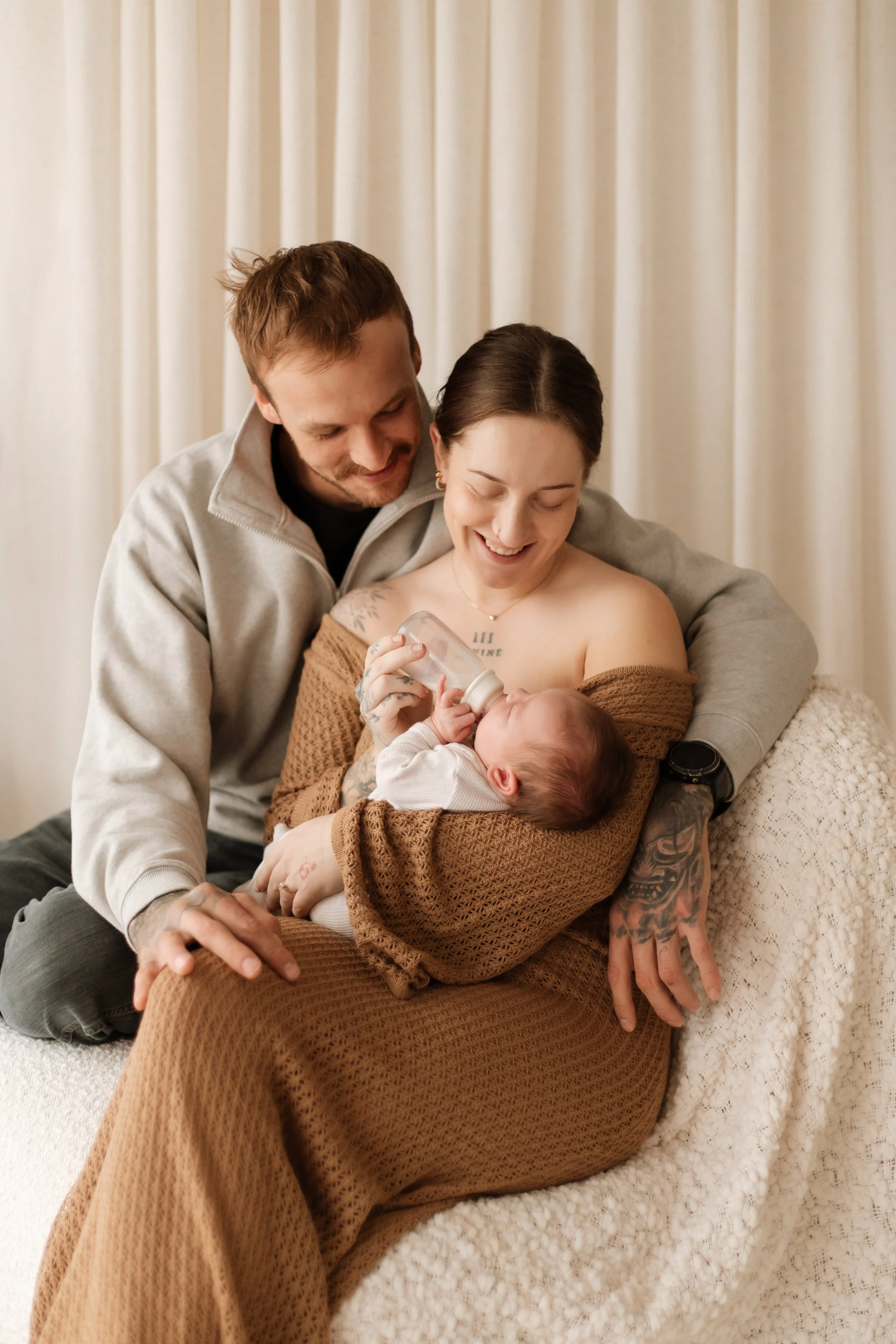 A happy family consisting of a woman, man, and newborn baby in a cozy setting with cream-colored curtains in the background. The woman is feeding the baby with a bottle, and both adults are smiling and looking at the infant.