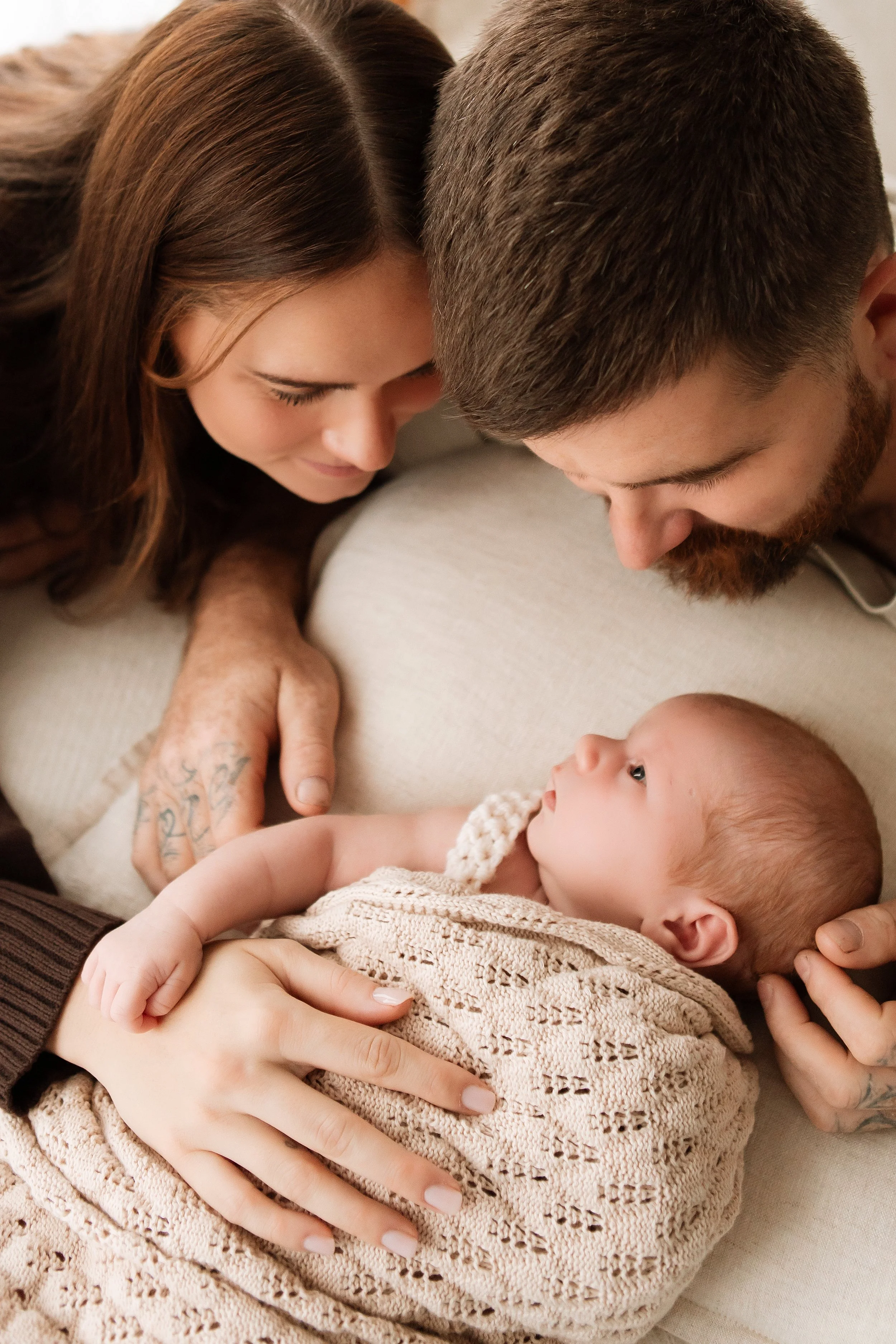 A woman, a man, and a baby are lying close together on a bed, with their heads touching. The woman has brown hair, and the man has short brown hair and a beard. They are looking lovingly at the baby, who is swaddled in a beige knitted blanket. The sc