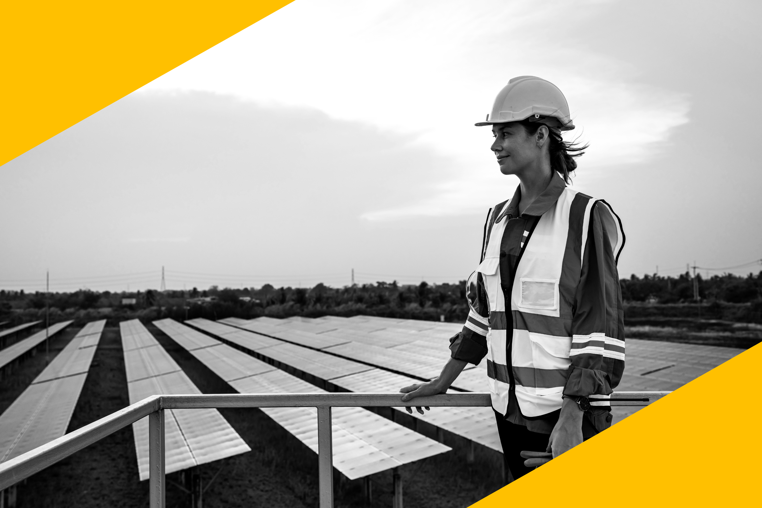 Eine Frauen in Bauarbeiterkleidung steht auf einer Plattform vor einer Solaranlage im Freien, im schwarz-weißen Bild. Sie trägt einen Schutzhelm und eine reflektierende Jacke, blickt nach rechts und hält sich an einem Geländer fest.