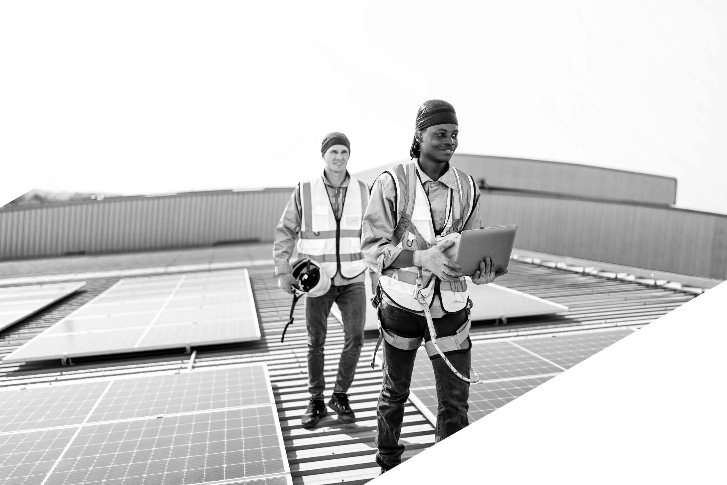 Zwei Bauarbeiterinnen auf einem Dach mit Solarmodulen, eine hält ein Tablet, die andere einen Helm, beide in Sicherheitskleidung