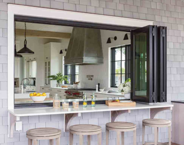 A kitchen viewed through a large pass-through window with a countertop and four bar stools outside. The inside features a range hood, windows, and kitchen counters with plants and utensils.
