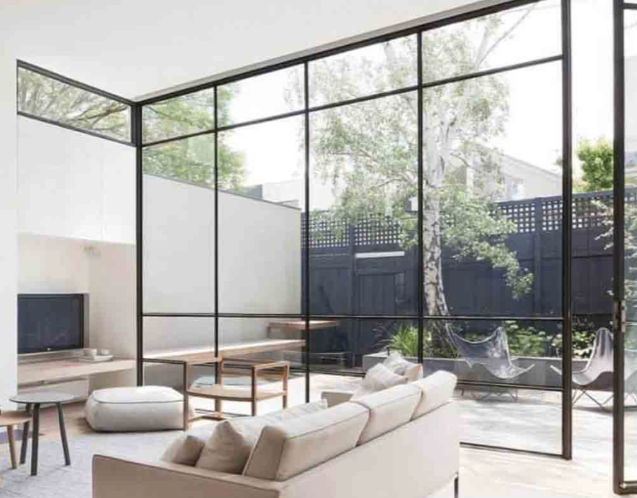 Modern living room with large floor-to-ceiling glass walls, outdoor patio with trees, outdoor chairs, and minimalist interior furniture.