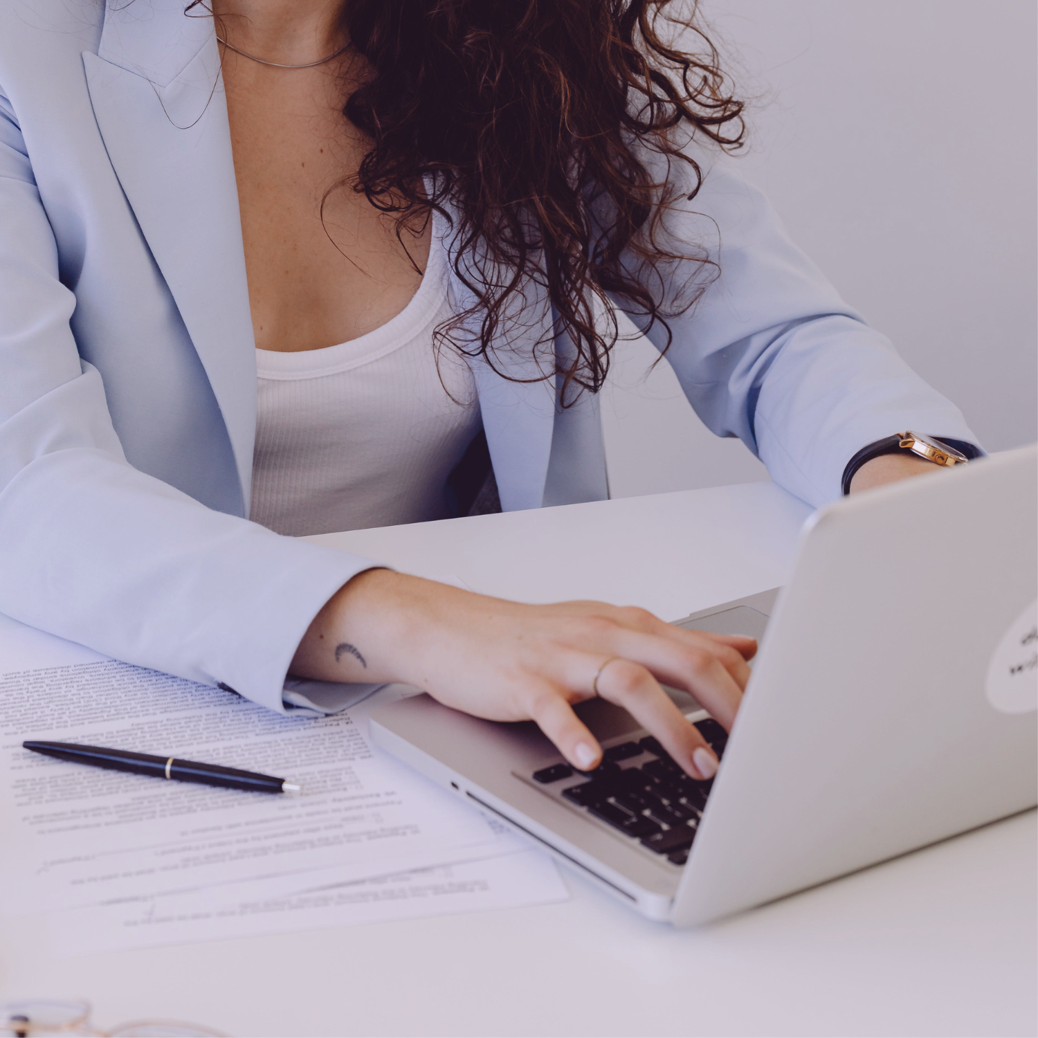 Woman with curly hair wearing a light blue blazer working on a silver laptop at a white desk, with documents and a pen nearby.