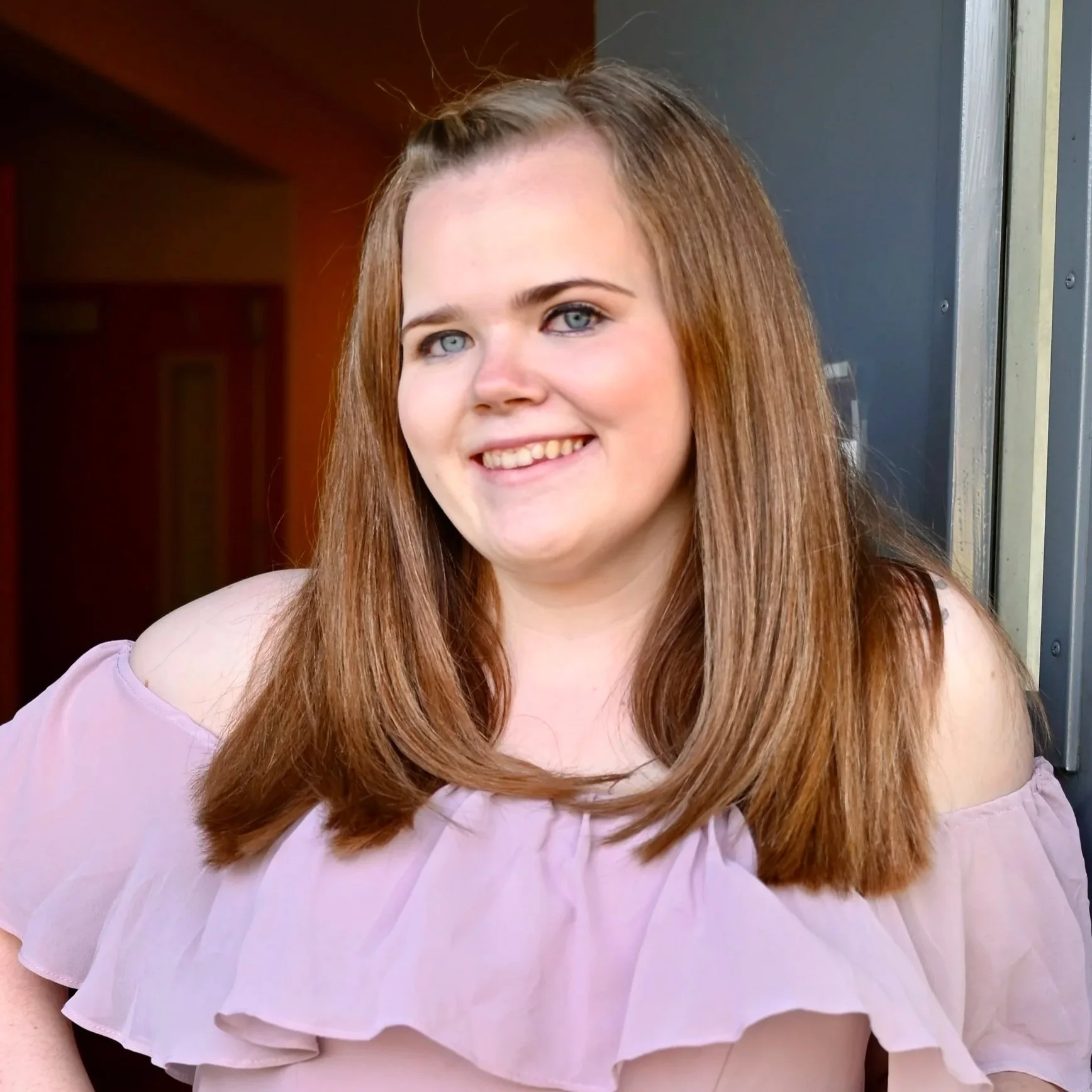 A young woman with long reddish-brown hair, blue eyes, and fair skin, smiling while standing outdoors near a building.