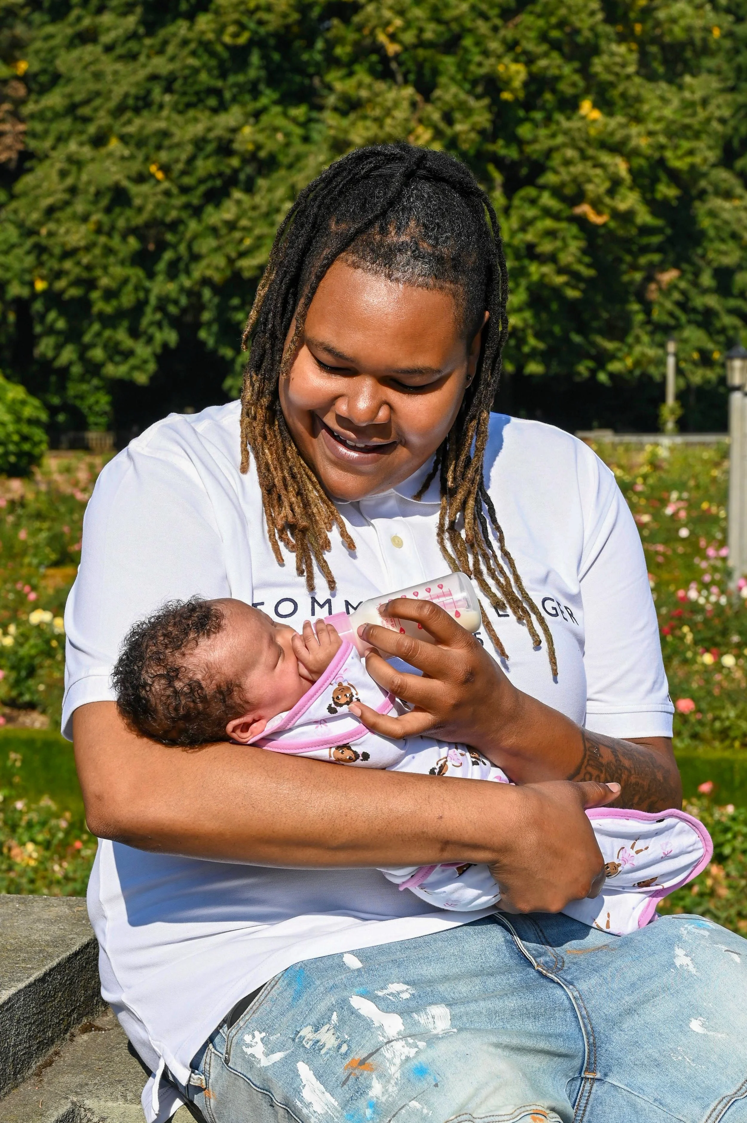 A woman with dreadlocks feeding a baby with a bottle in a park.