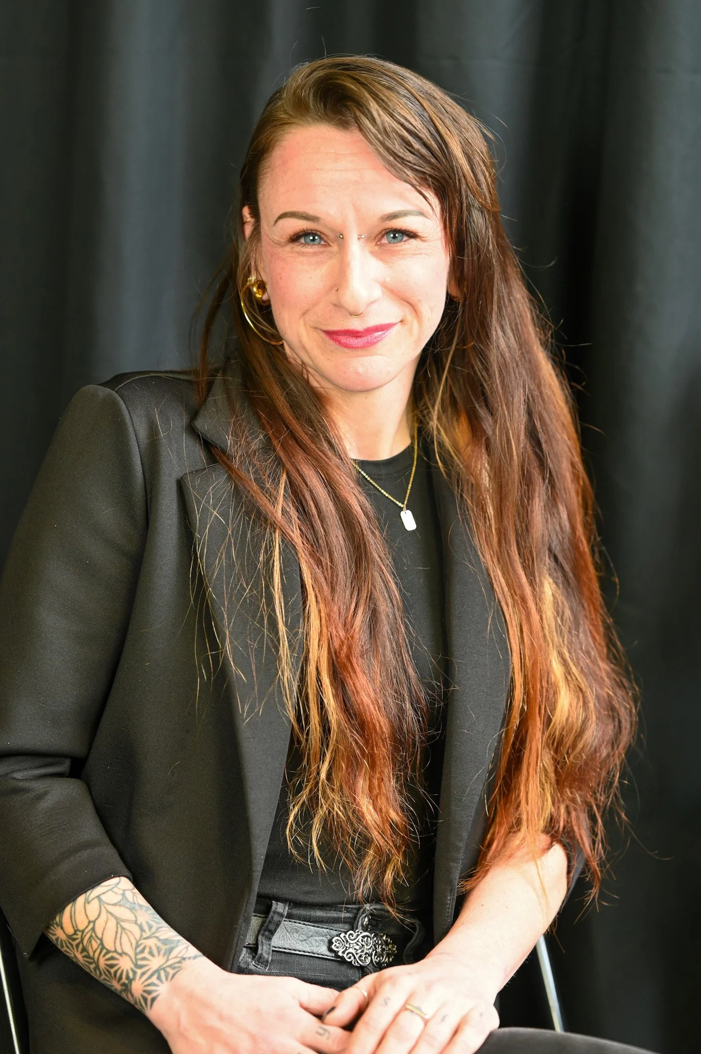 A woman with long reddish-brown hair, wearing a black blazer, gold hoop earrings, and a black top, sitting against a black background.