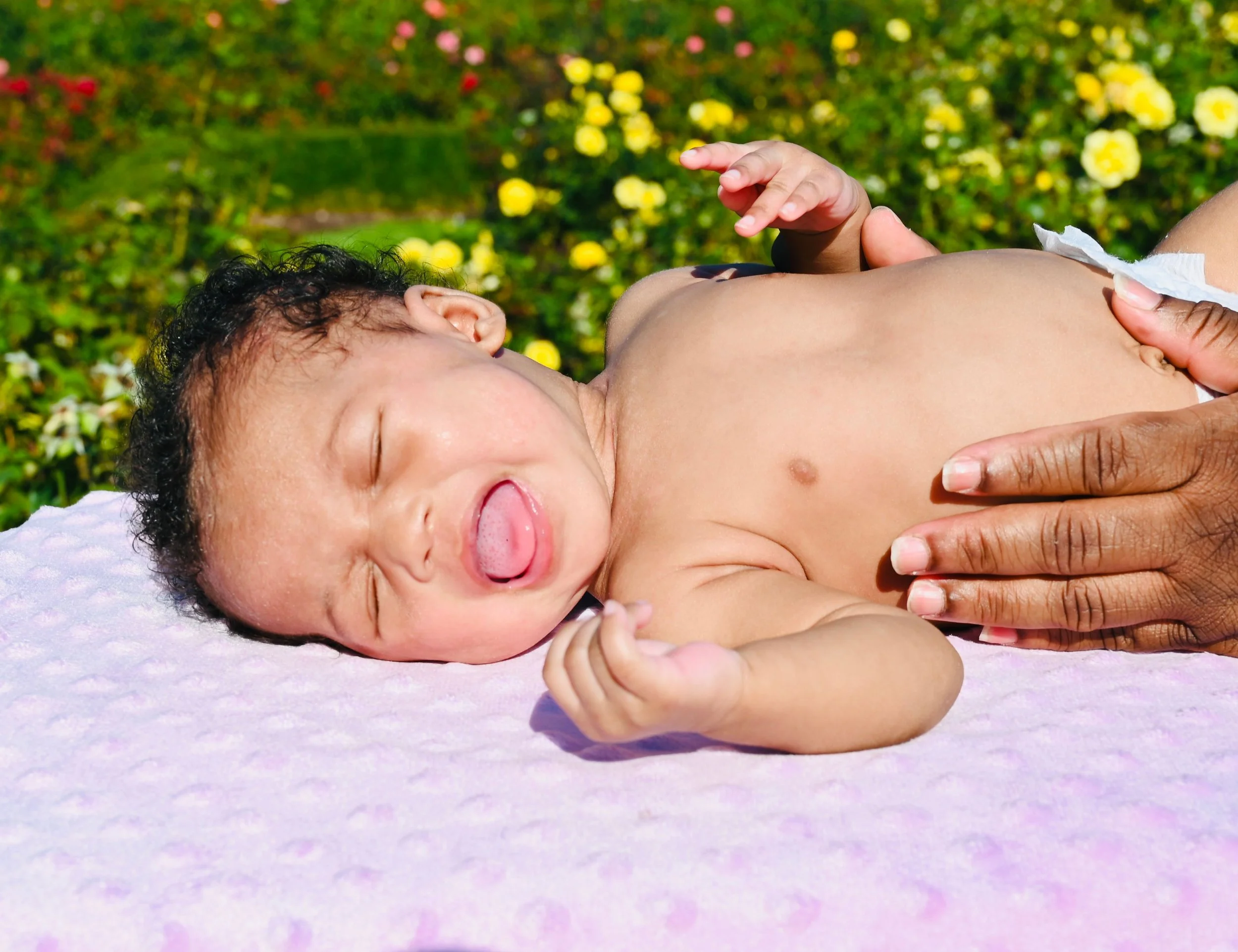 A baby lying on a pink blanket outdoors with a field of yellow and white flowers in the background, sticking out their tongue.