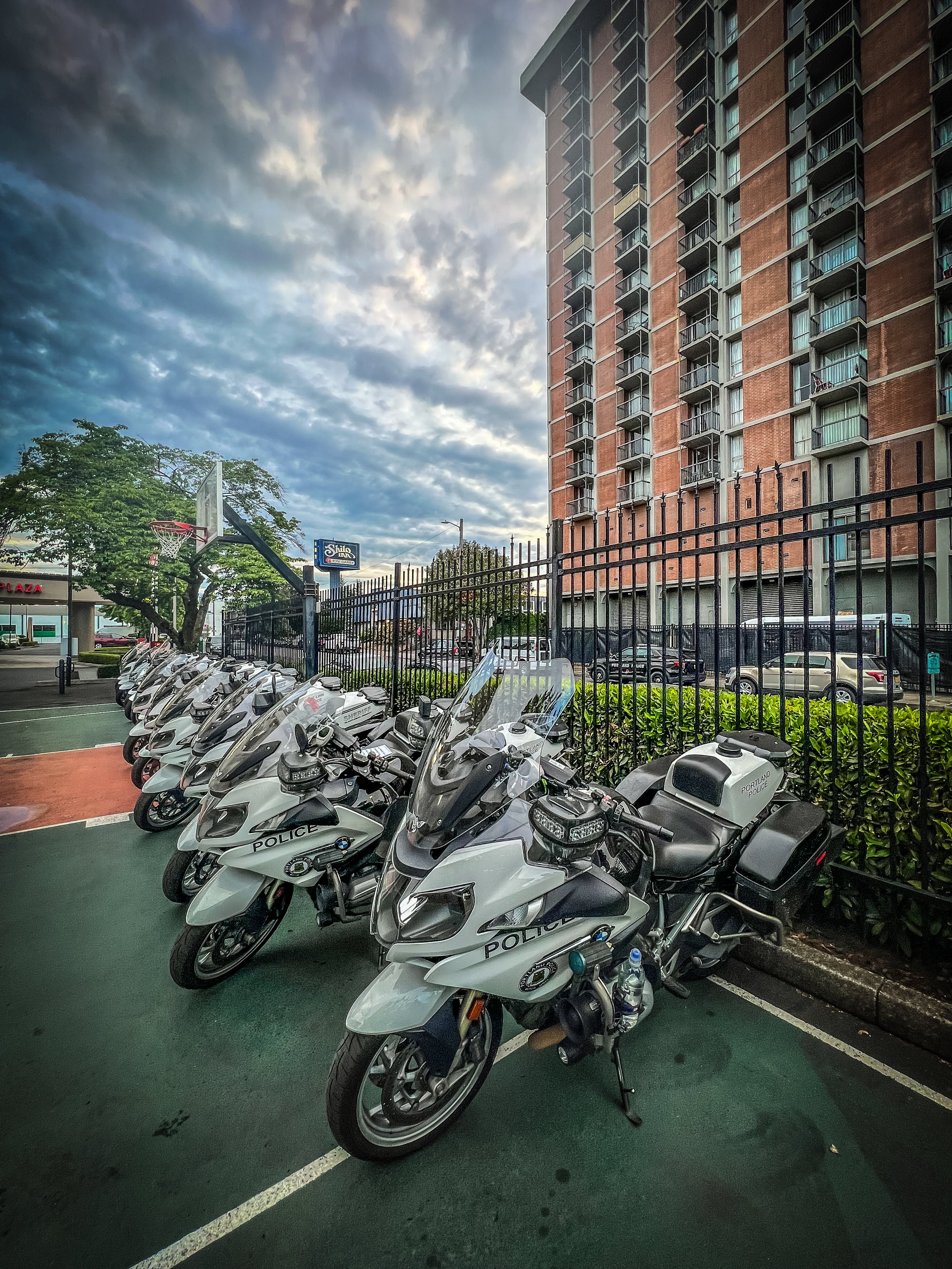 Multiple police motorcycles parked in a row on a parking lot, with a tall multi-story apartment building in the background and cloudy sky overhead.
