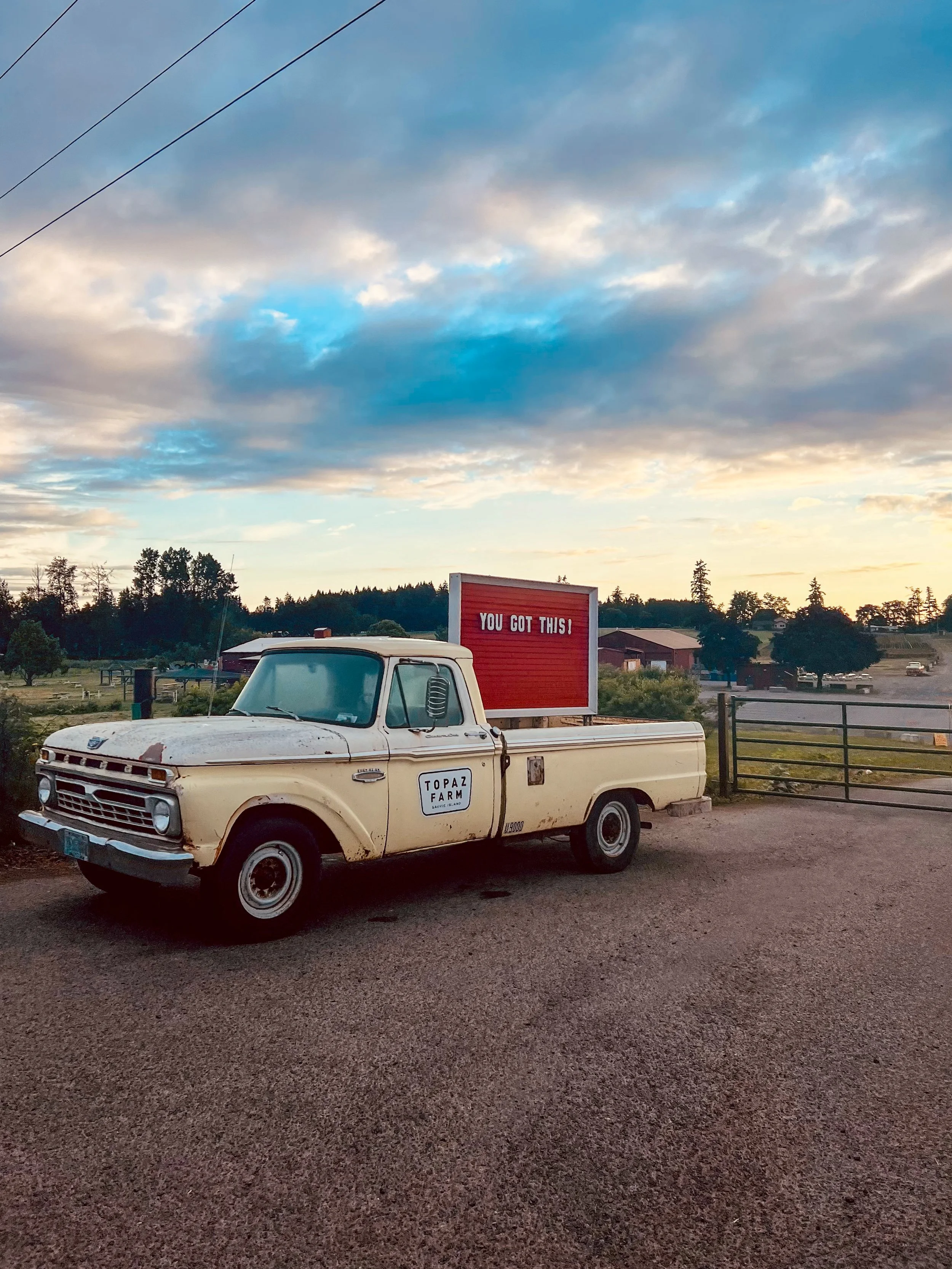 An old beige pickup truck with a 'TOPAZ FARM' sign on the door, parked on gravel near a fence. There is a red billboard on the truck's bed with white text that reads 'YOU GOT THIS!'. The background features a rural landscape with trees, buildings, an