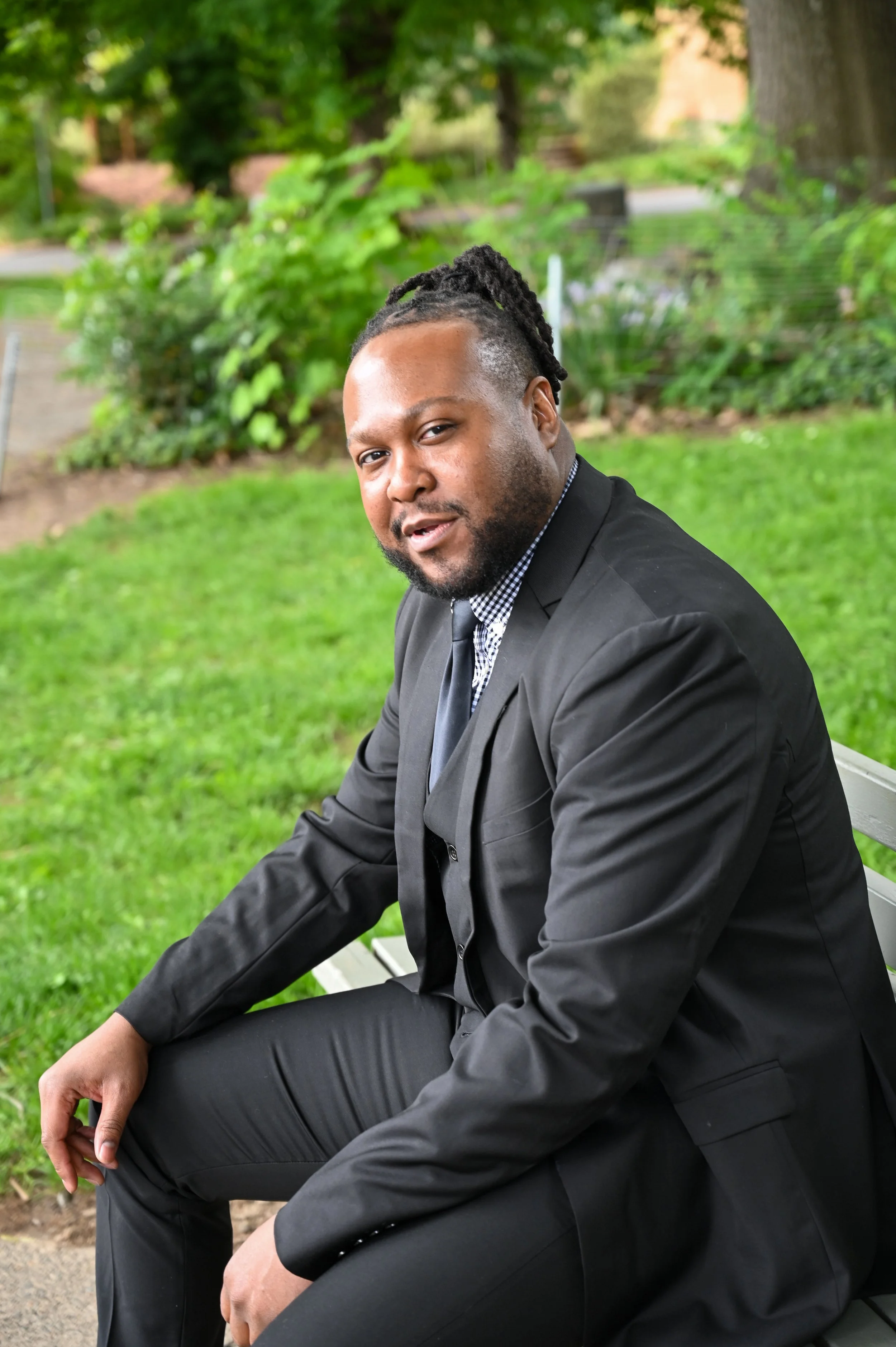 A man in a black suit and checkered shirt sitting on a park bench outdoors, surrounded by green grass and trees.