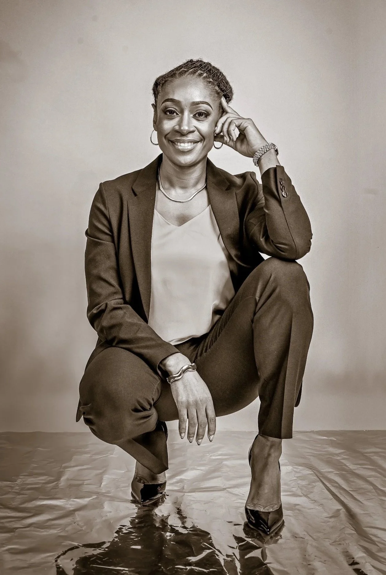 Rashida Saunders in a business suit squatting with one knee raised, smiling, with her hand resting on her temple, in a studio setting.