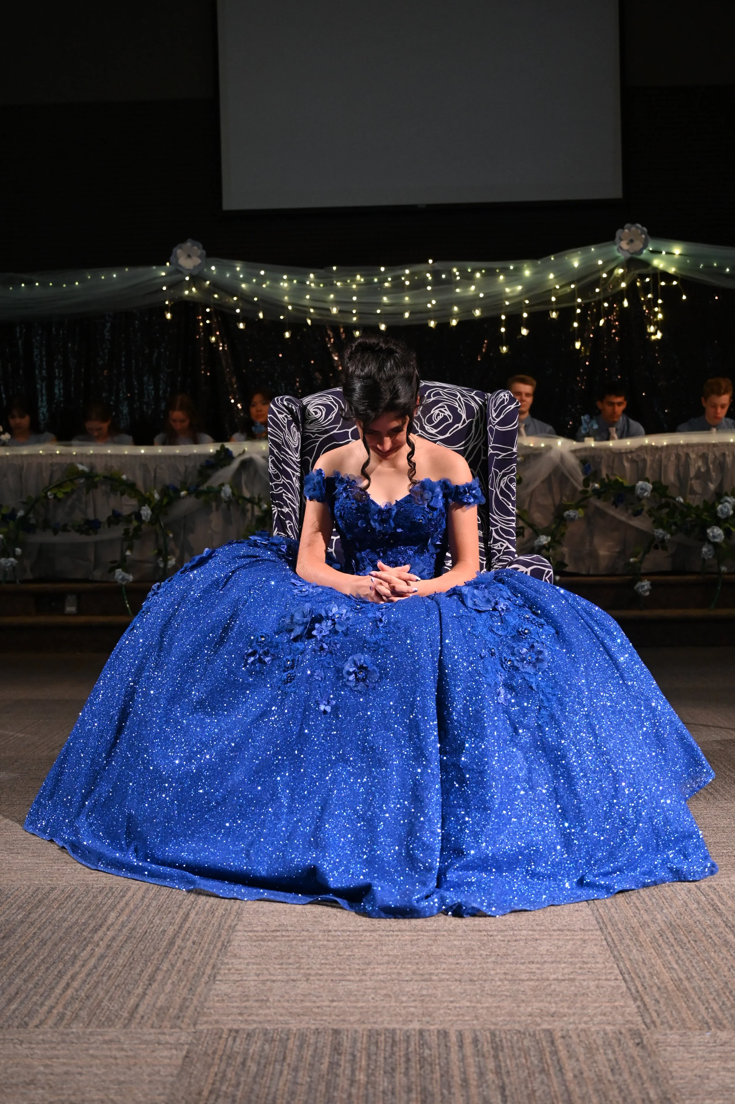 A girl sitting on a black and white patterned chair wearing a sparkly blue dress with floral details at a formal event.