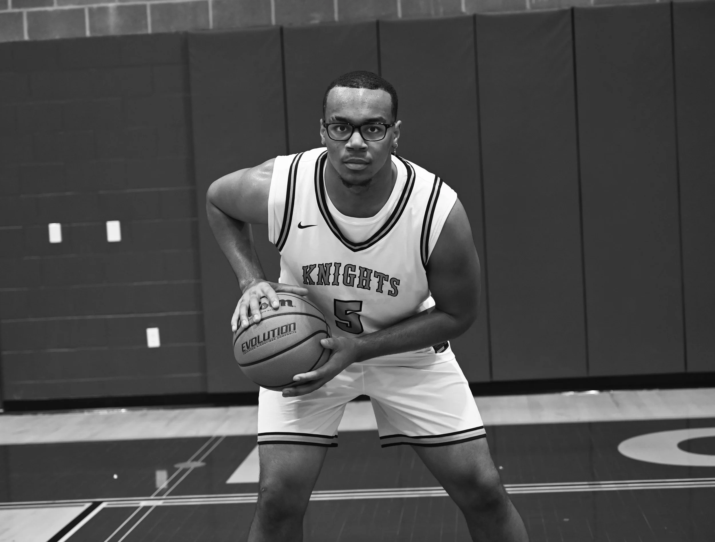 A young man with glasses in a basketball uniform holding a basketball on a basketball court.
