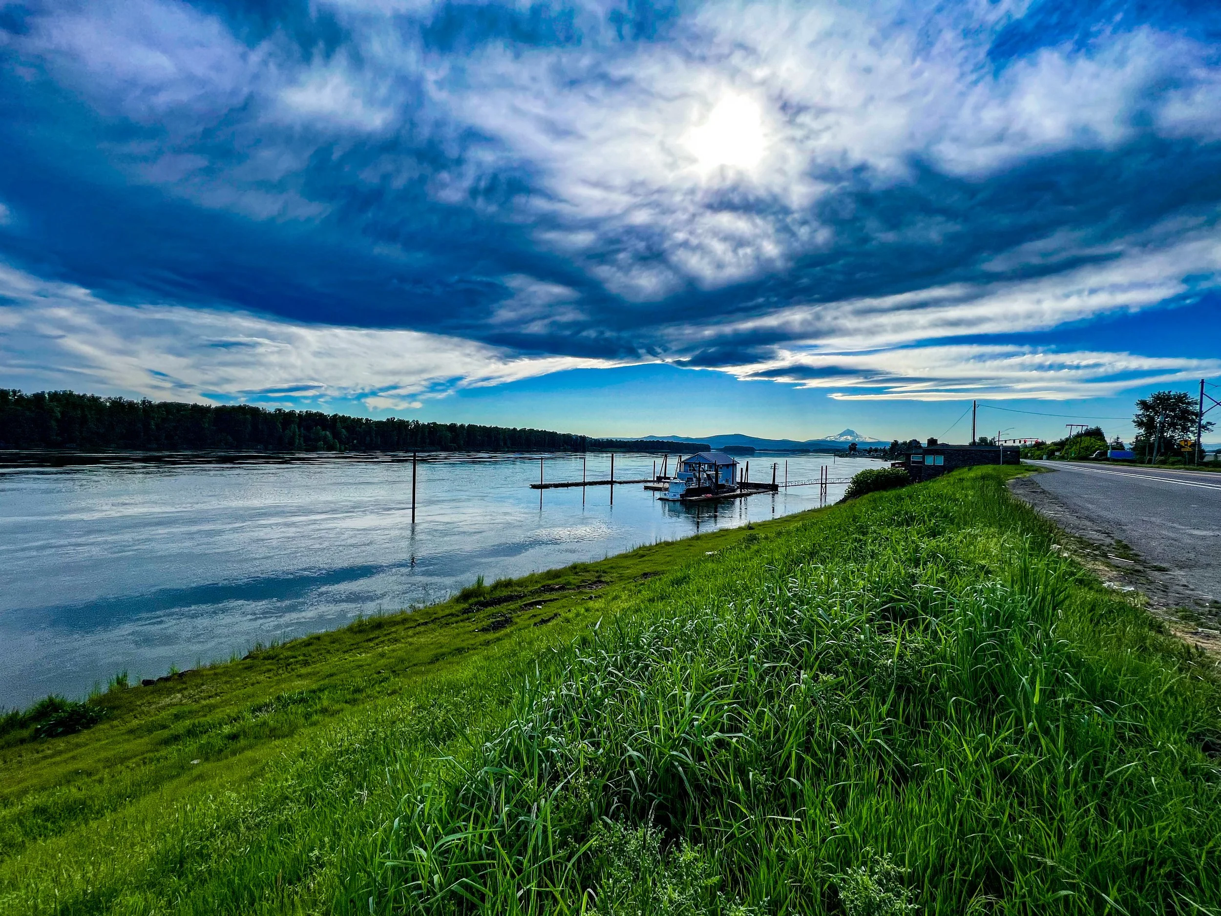 A river scene with calm water, greenery along the shoreline, a dock with boats, a road on the right, and a partly cloudy sky with the sun shining through.