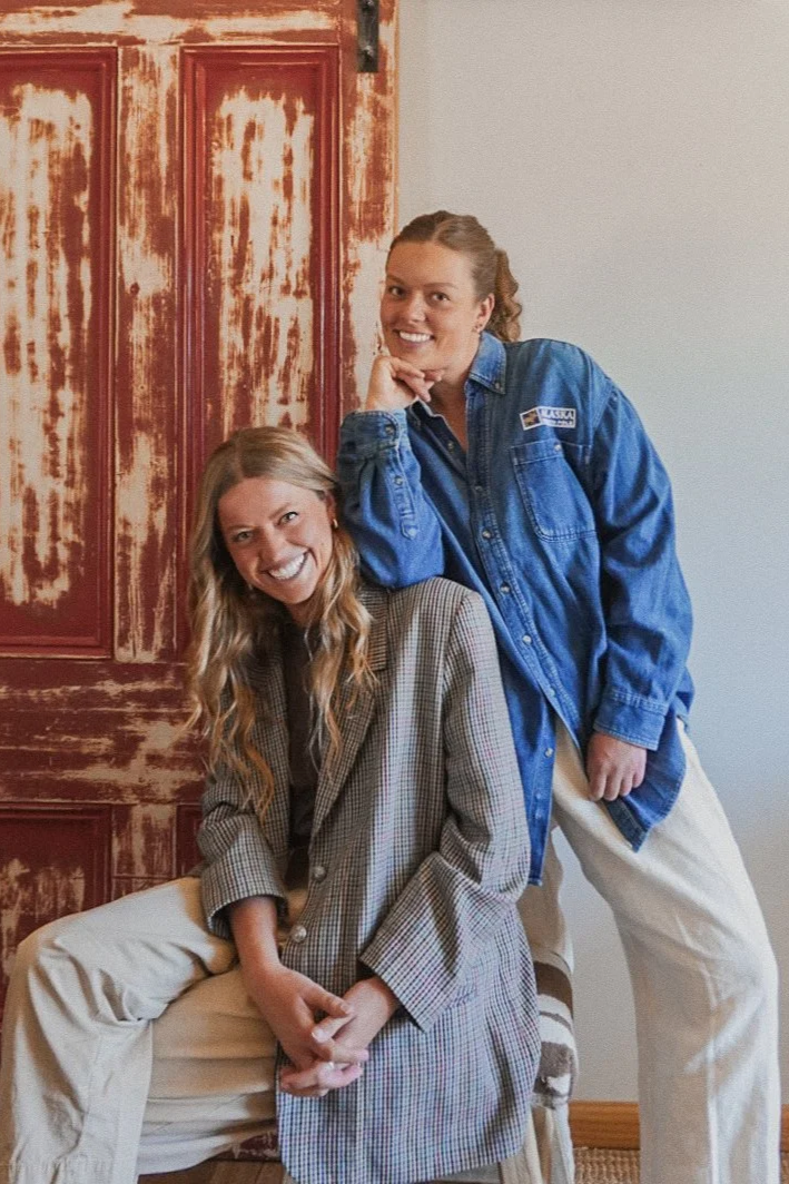 Two women posing together indoors. One woman is sitting on a chair, wearing a plaid blazer and beige pants, smiling. The other woman stands behind her, leaning on her shoulder, wearing a blue denim jacket and white pants, also smiling. In the background, there is a rustic, red, distressed wooden door.
