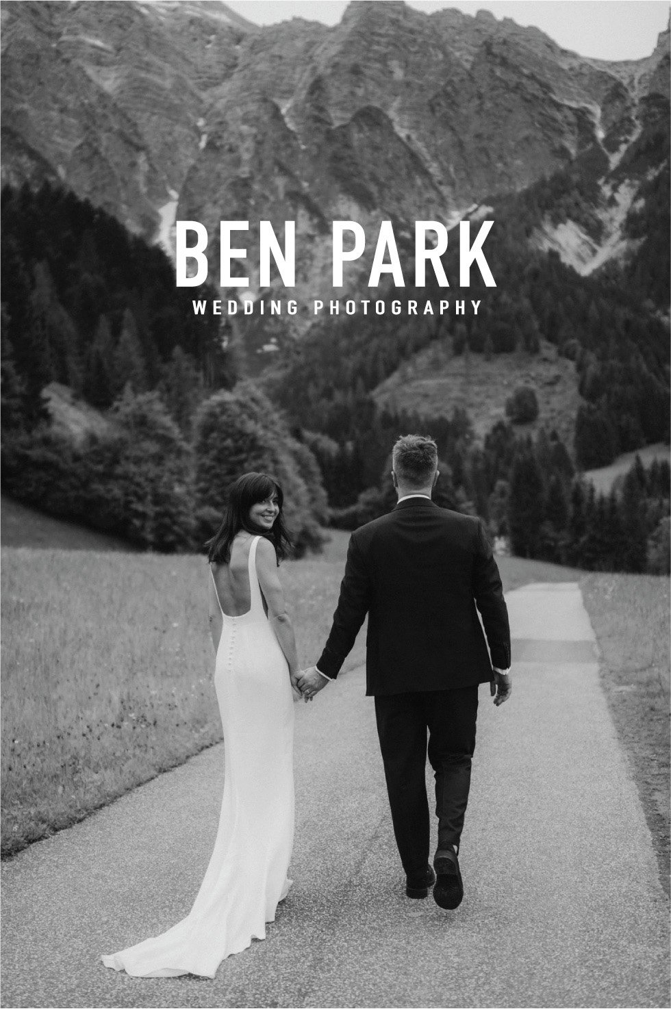 Black and white photo of a bride and groom holding hands, walking down a road with mountains in the background, for wedding photography by Ben Park.